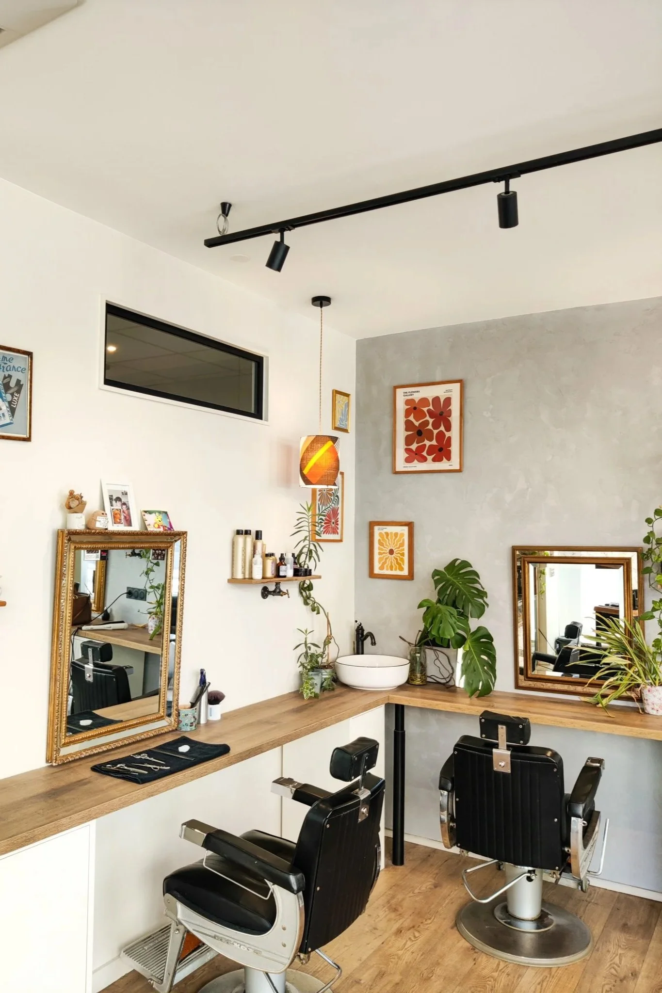 Intérieur d'un salon de coiffure avec deux fauteuils de barbier, deux miroirs, des plantes vertes, des produits capillaires, une lampe suspendue, des cadres avec des œuvres d'art sur un mur gris, et un éclairage moderne.