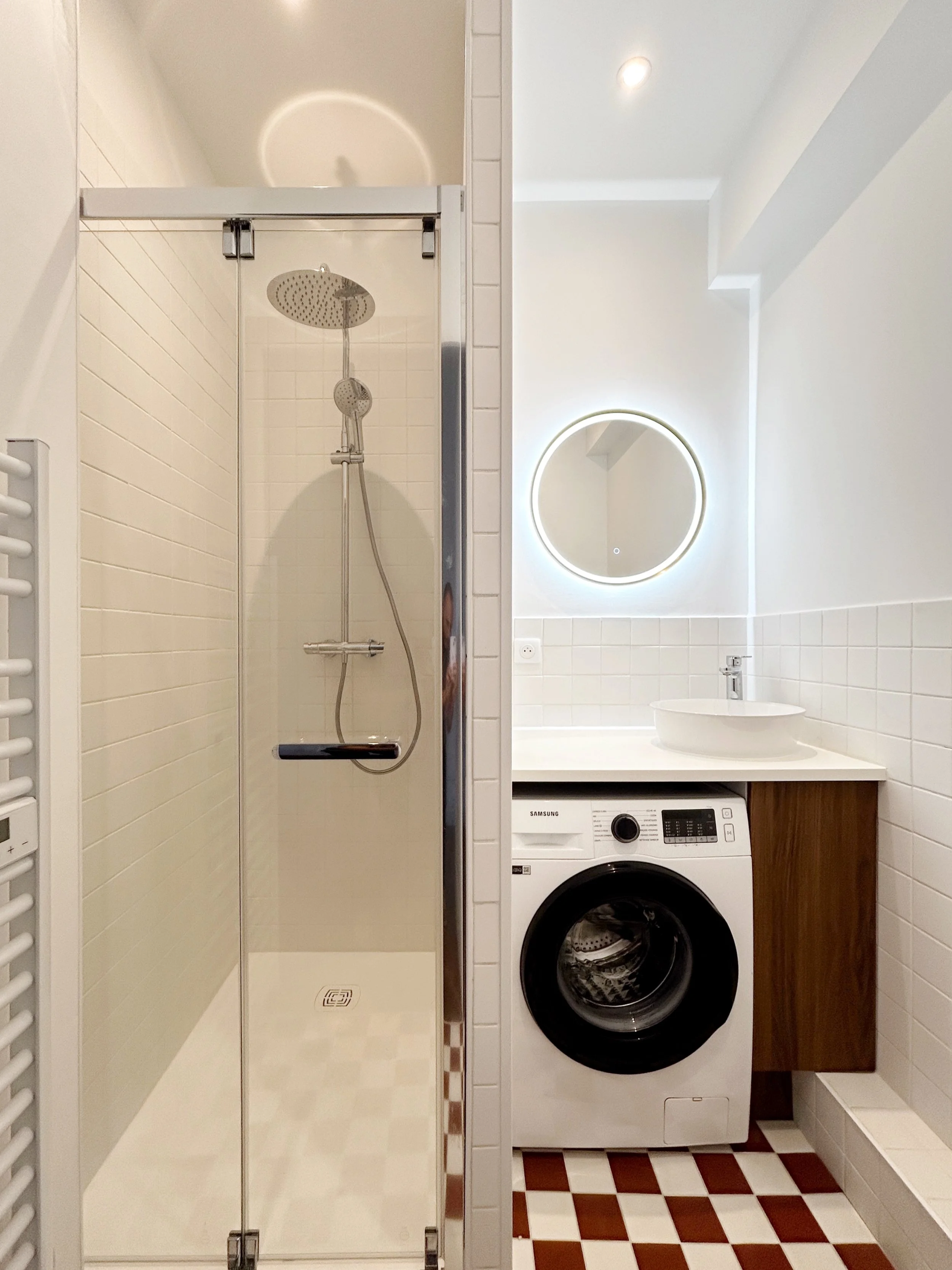 Une salle de bain comprenant une cabine de douche avec paroi en verre, une machine à laver, un lavabo avec un miroir lumineux, un sol en carreaux bicolores, des murs en carreaux clairs, et un chauffage rayonnant blanc.