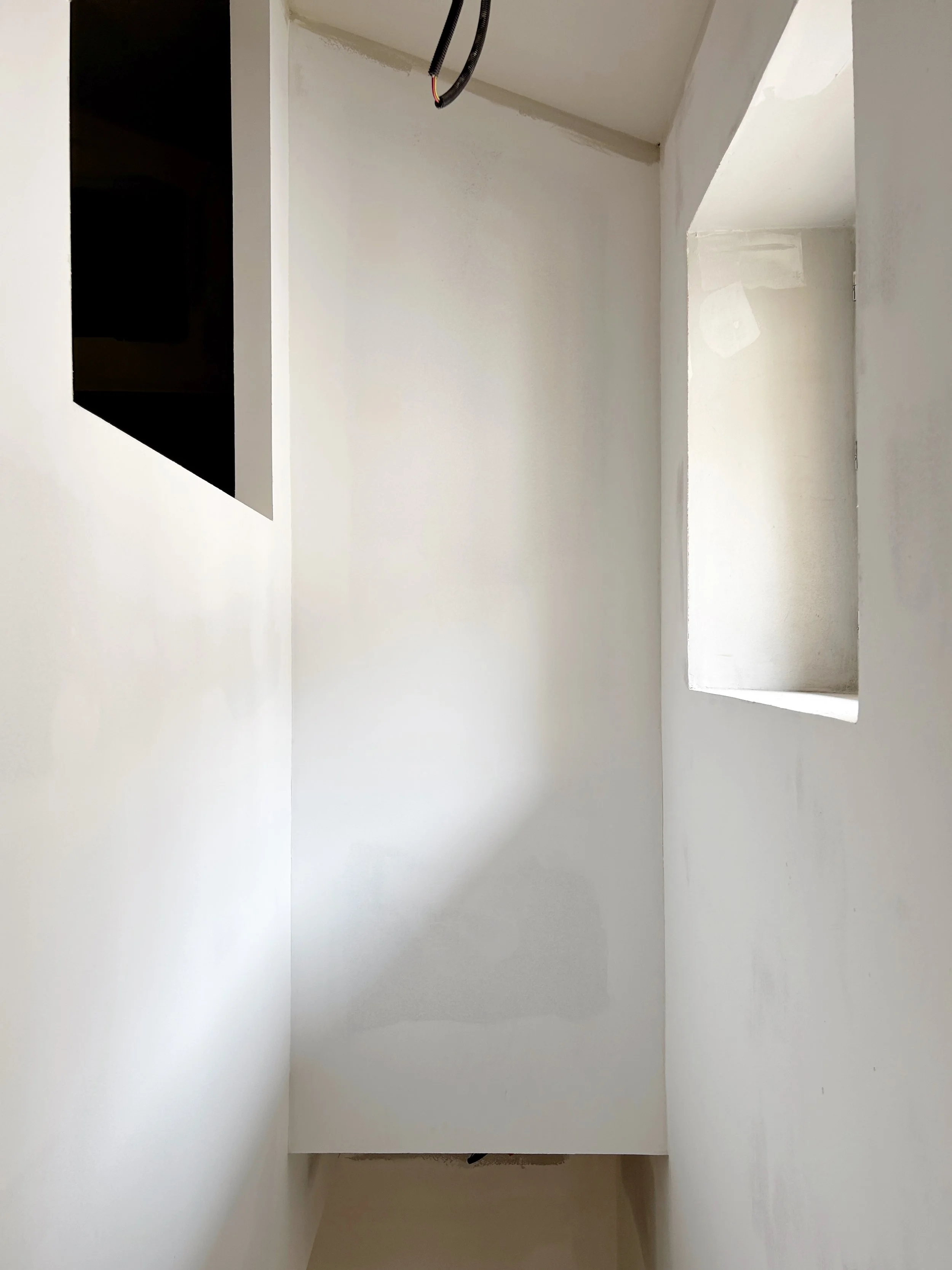 Une pièce en cours de rénovation avec des murs peints en blanc, un grand espace ouvert, un petit placard ou une alcôve, et des câbles suspendus au plafond.