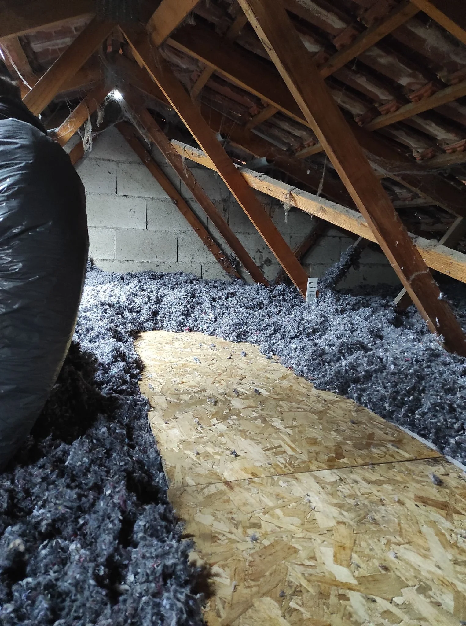 Intérieur d'une pièce isolée avec du rembourrage en laine de roche, un panneau de contreplaqué posé par terre, et des poutres en bois au plafond.
