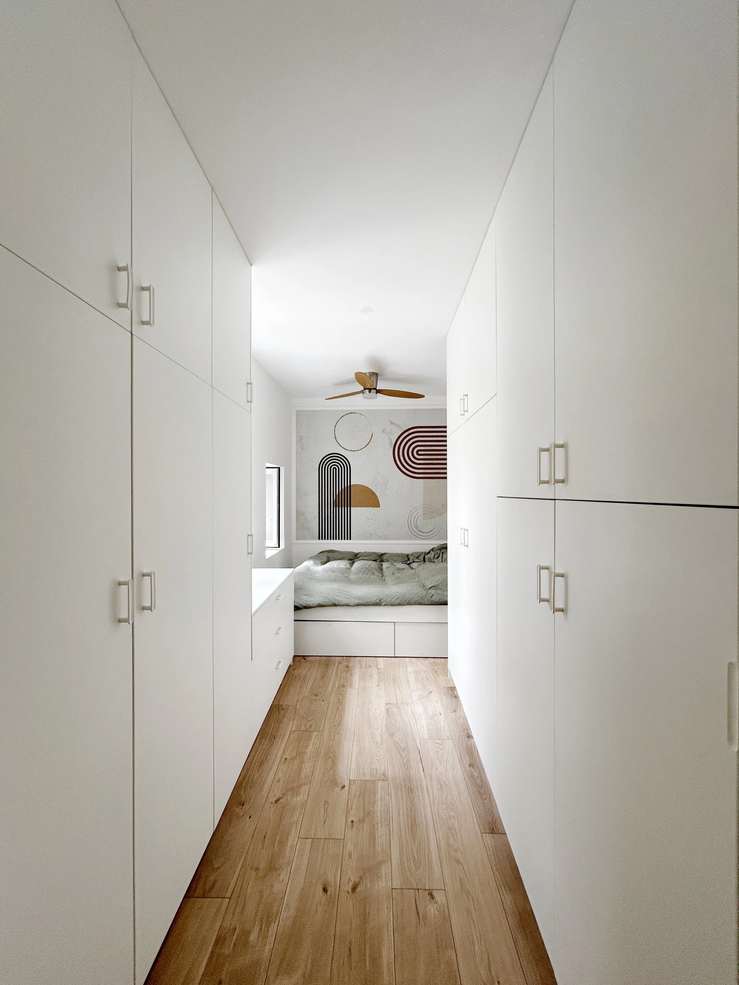 Une pièce hexagonale avec des armoires blanches sur les côtés et un lit avec des draps gris clair à l'extrémité, décoration abstraite sur le mur derrière le lit, ventilateur au plafond en bois.