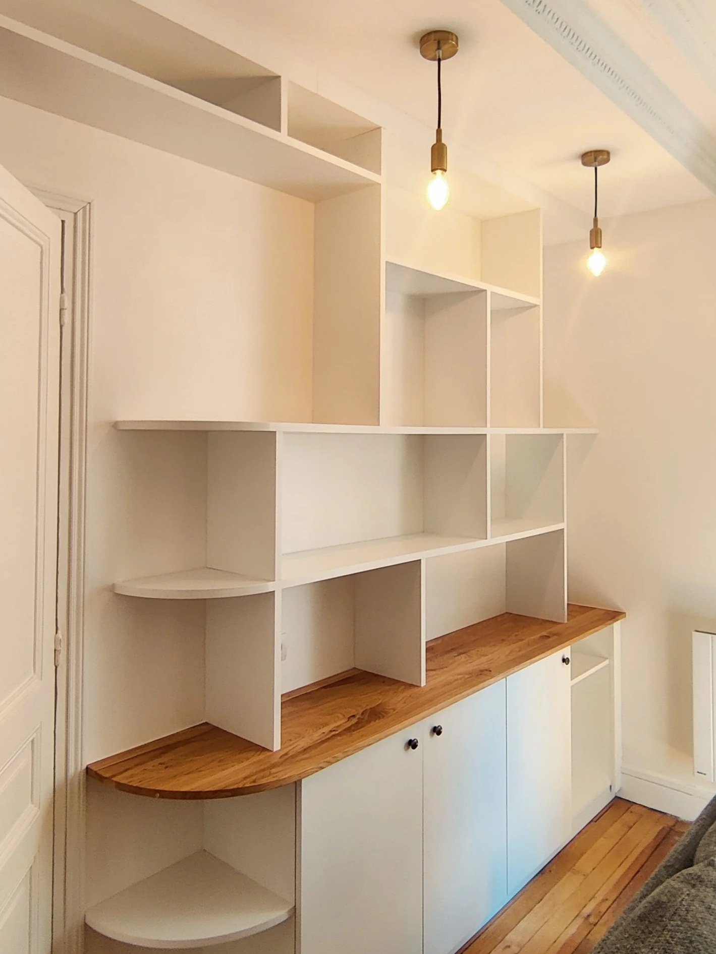 Un meuble blanc avec des étagères et un plan de travail en bois clair dans une pièce. Deux lampes suspendues lumineuses au plafond.