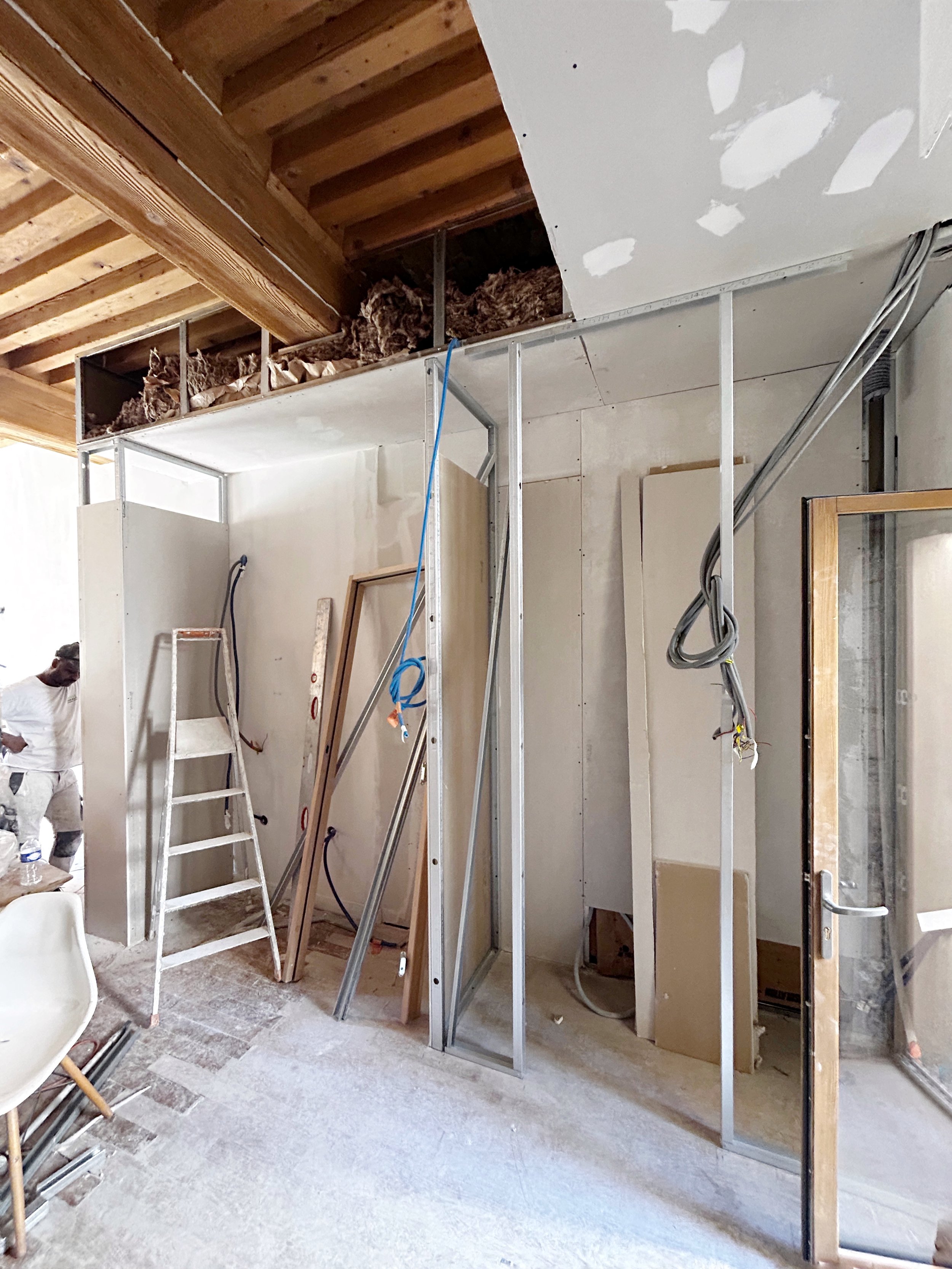 Travaux en cours dans une pièce en construction, avec des murs en placo, une échelle en métal, un mur partiellement monté, des câbles électriques apparents, une personne en arrière-plan, et un plafond en bois apportant une touche chaleureuse.