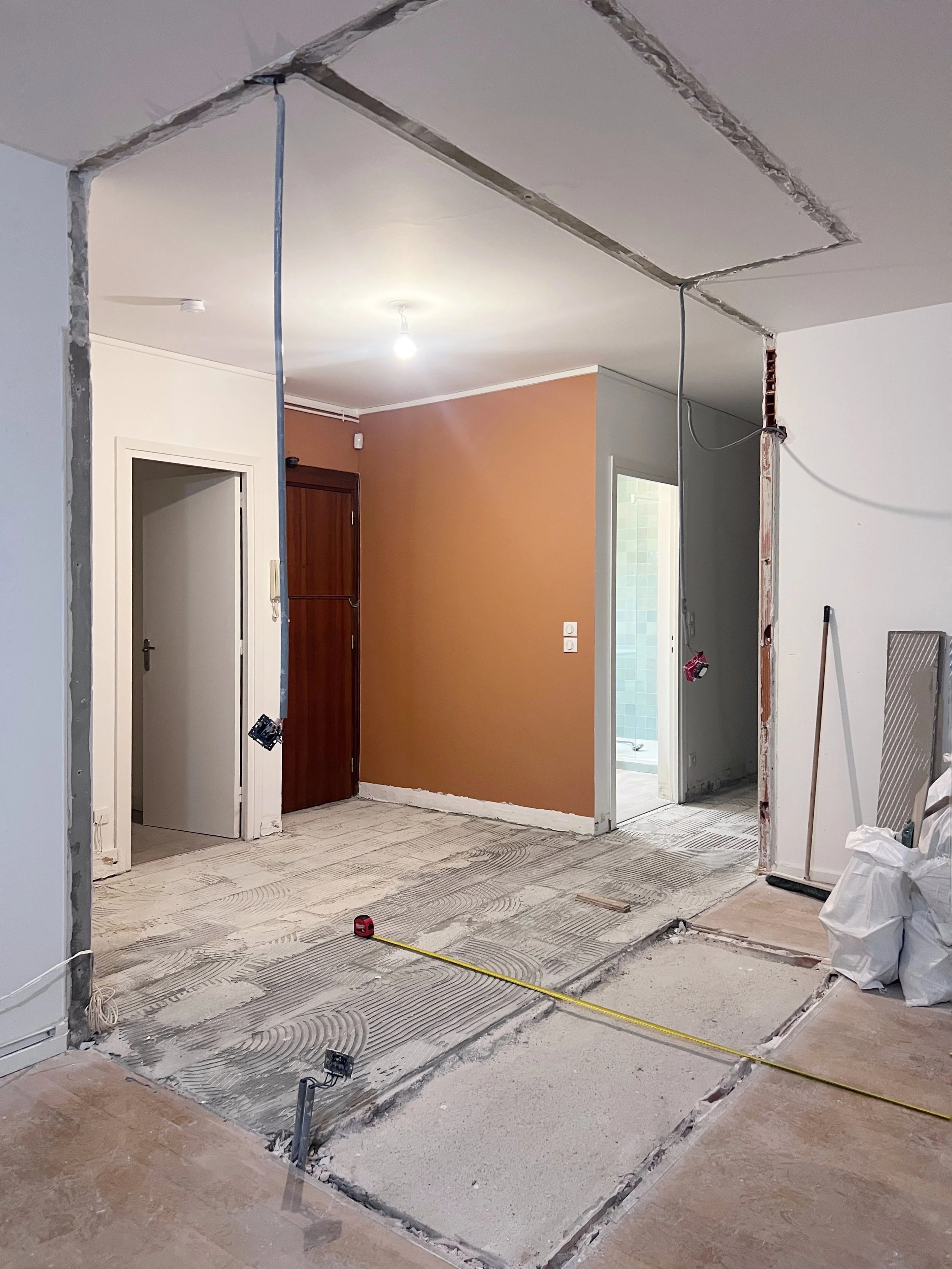 Travaux de rénovation dans une pièce où le sol est préparé avec des bandes de mousse isolante pour une nouvelle couche de revêtement. Les murs sont en cours de finition avec des câbles électriques visibles, et une porte mène à une autre pièce. Il y a des outils et matériaux de construction autour.
