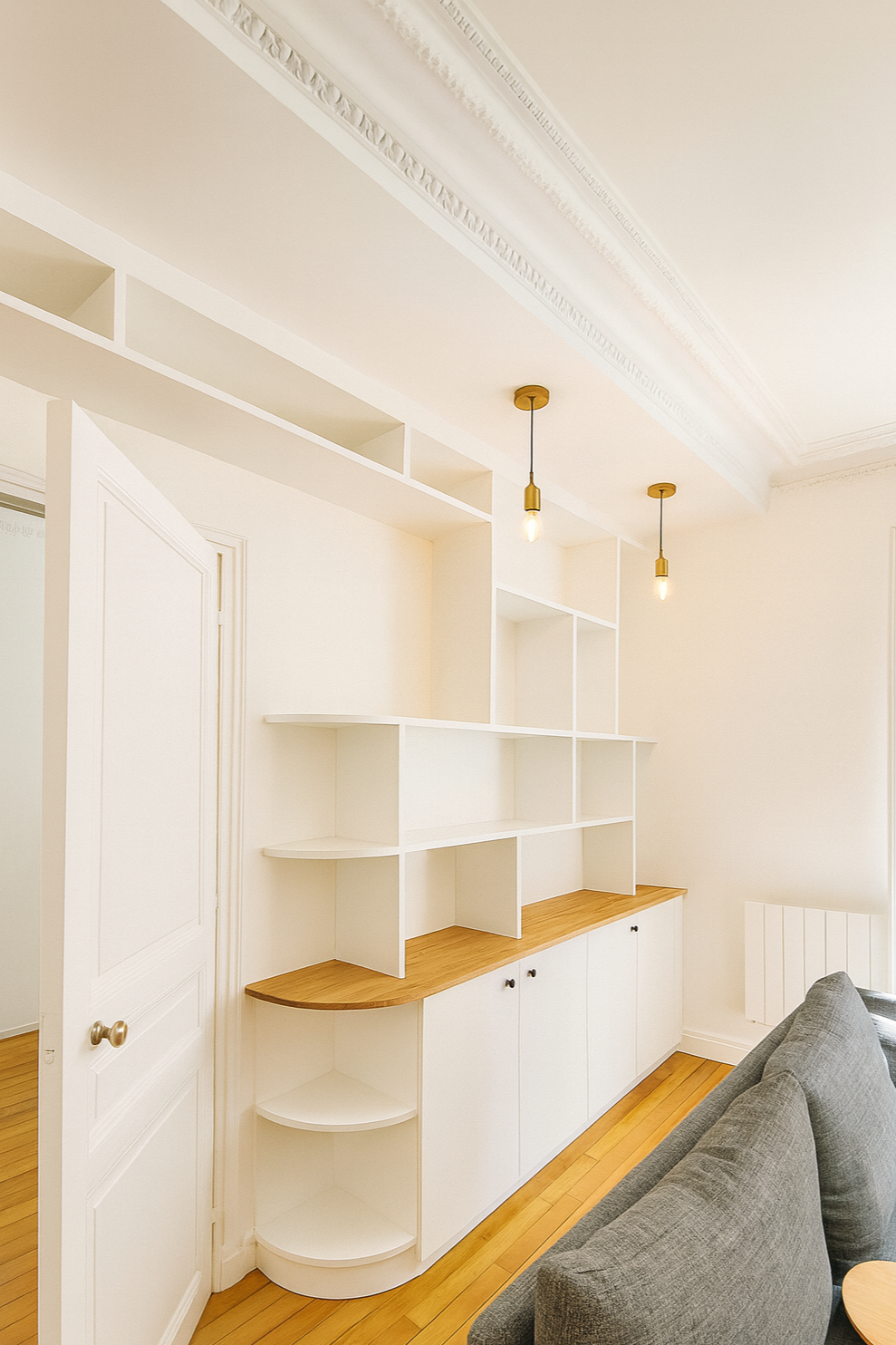 Pièce lumineuse avec un meuble blanc à étagères et une surface en bois, canapé gris, plafond orné de moulures, éclairage suspendu, murs blancs.