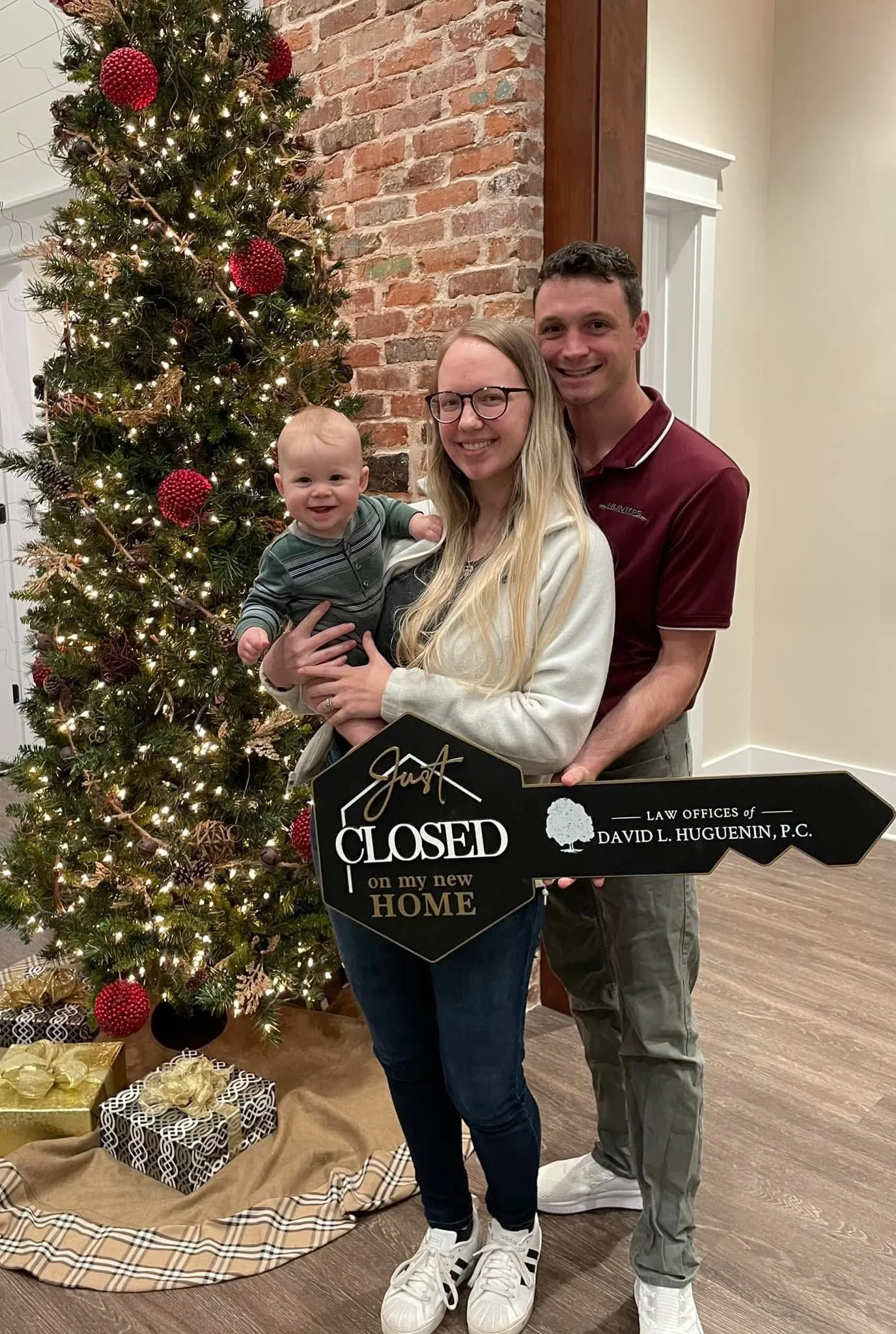 Family of three standing in front of Christmas tree holding a sign that says 'Just CLOSED on my new HOME' with presents underneath.