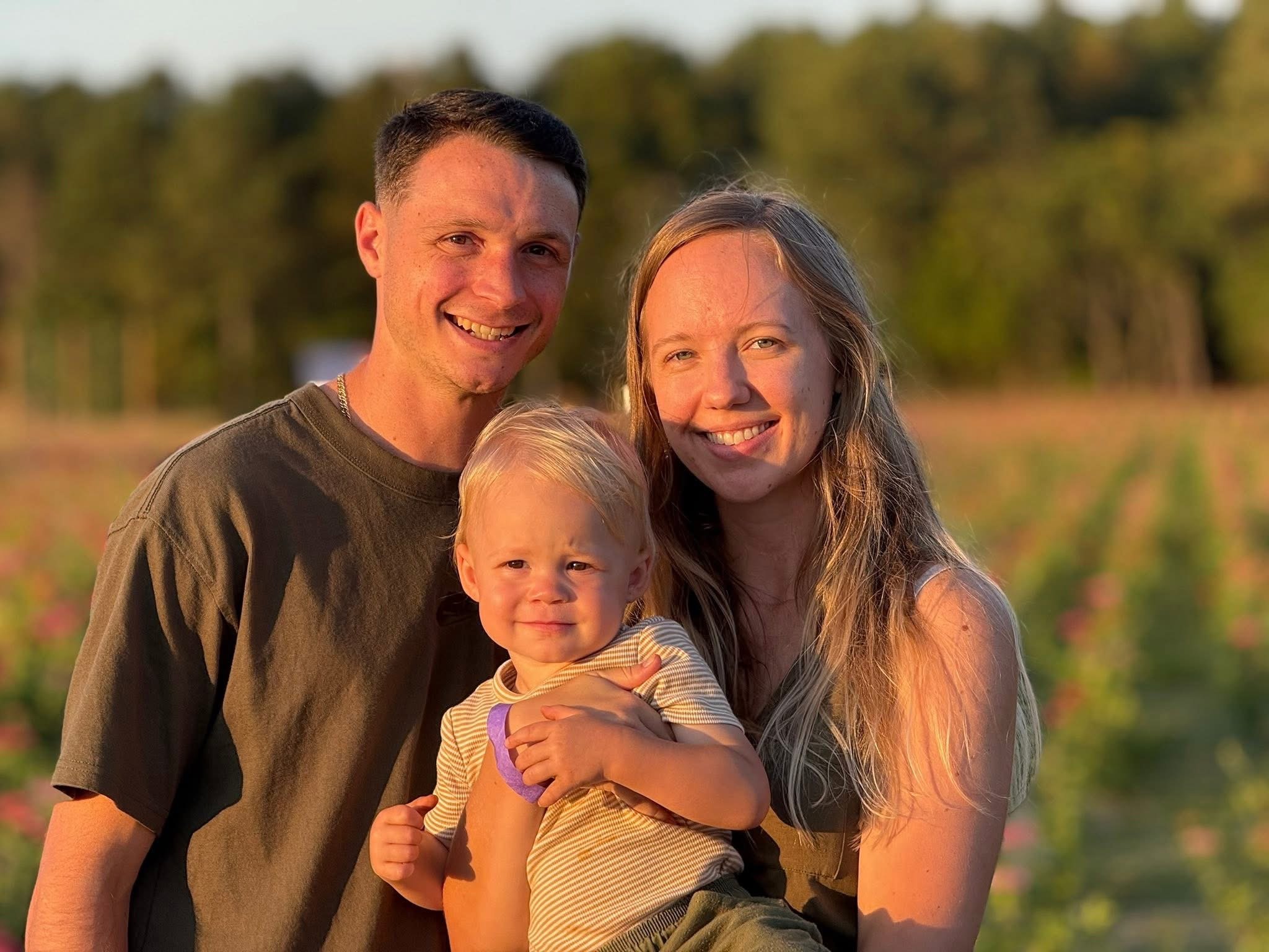 A happy family of three, a man, a woman, and a young child, smiling outdoors during sunset in a field with trees in the background.