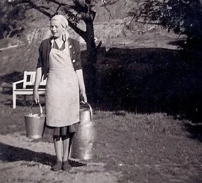 En kvinne står ute i naturen med to bøtter, iført apron og kledd i eldre tidstypisk klær.