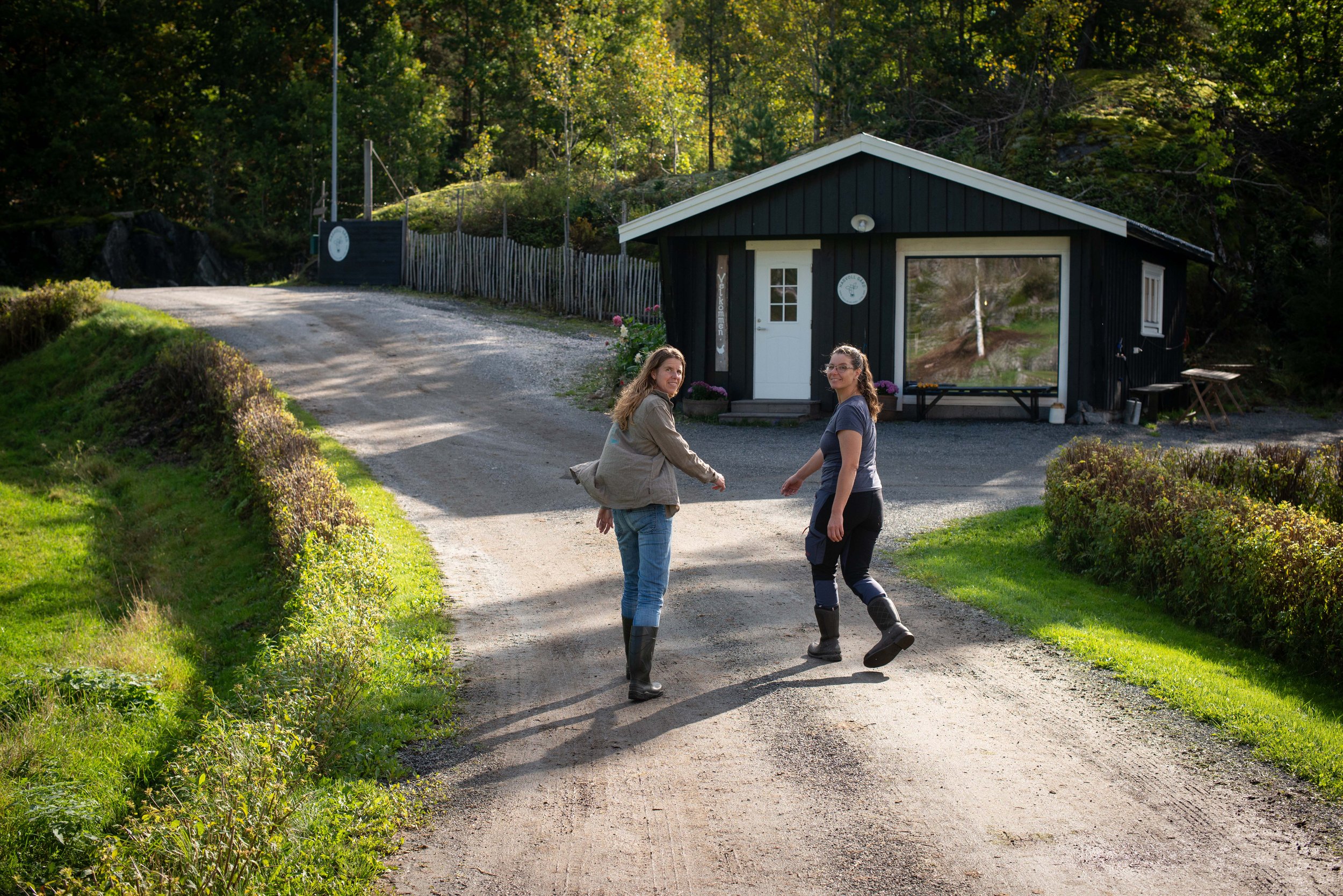 To kvinner går på en grusvei foran en liten svart hytte med hvite detaljer, omgitt av grønt landskap og trær.