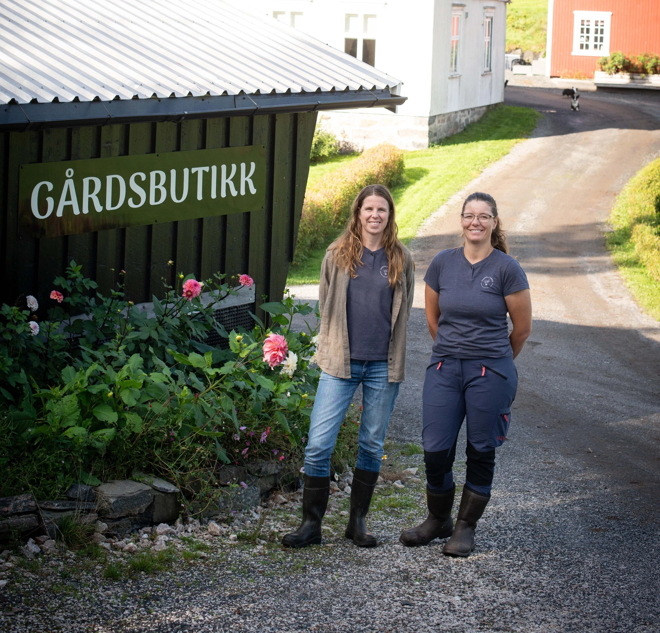 To kvinner står ved en jordbutikk, smilende, på en grusvei foran en svart bygning med et skilt som sier 'GÅRDSBUTIKK'.