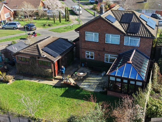 A two-story red brick house with solar panels on the roof, a backyard with a patio, a small storage shed, and a glass-enclosed sunroom. A person is standing near the shed, and cars are parked on the street nearby.