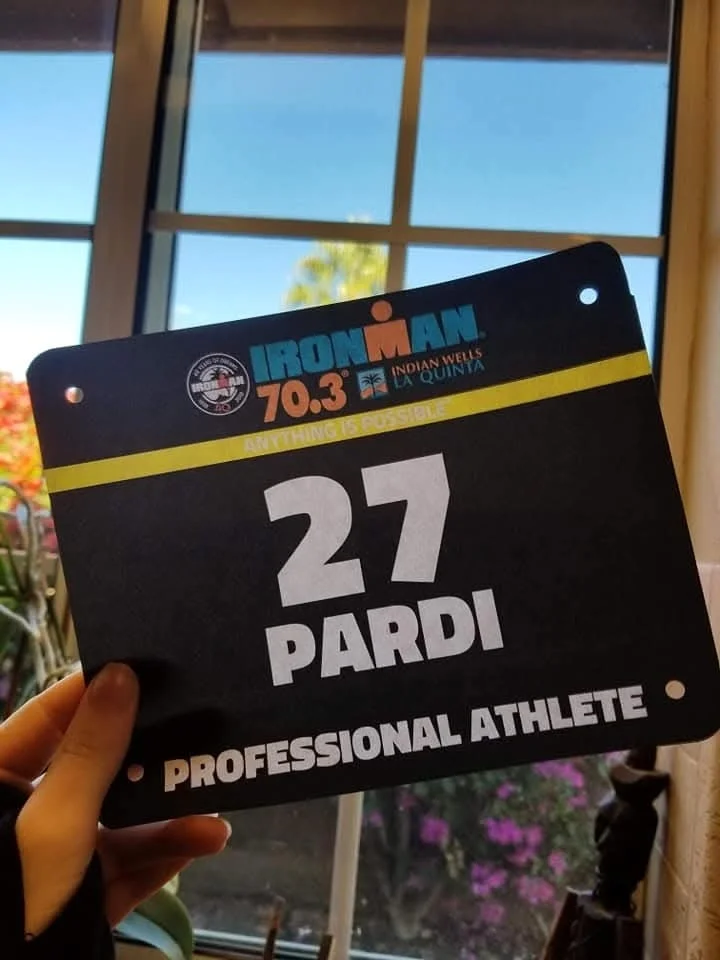 A race bib with the name Pardi, the number 27, and the word Professional Athlete. The bib is for Ironman 70.3 in Indian Wells, La Quinta, with the slogan "Anything is Possible." It is held outdoors near a window with a blue sky and some flowers visible in the background.