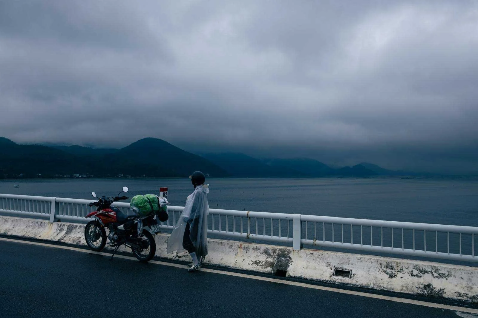 A person in a raincoat walking near a motorcycle on a bridge over a body of water, with mountains in the background under a cloudy sky.