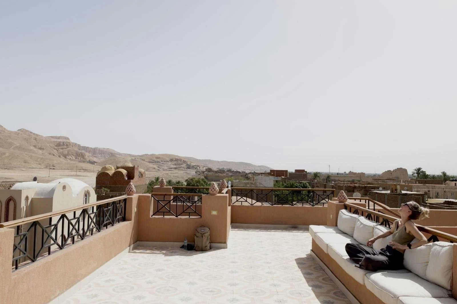 A woman sitting on a white outdoor sofa on a terrace under a bright sky with a desert landscape in the background.