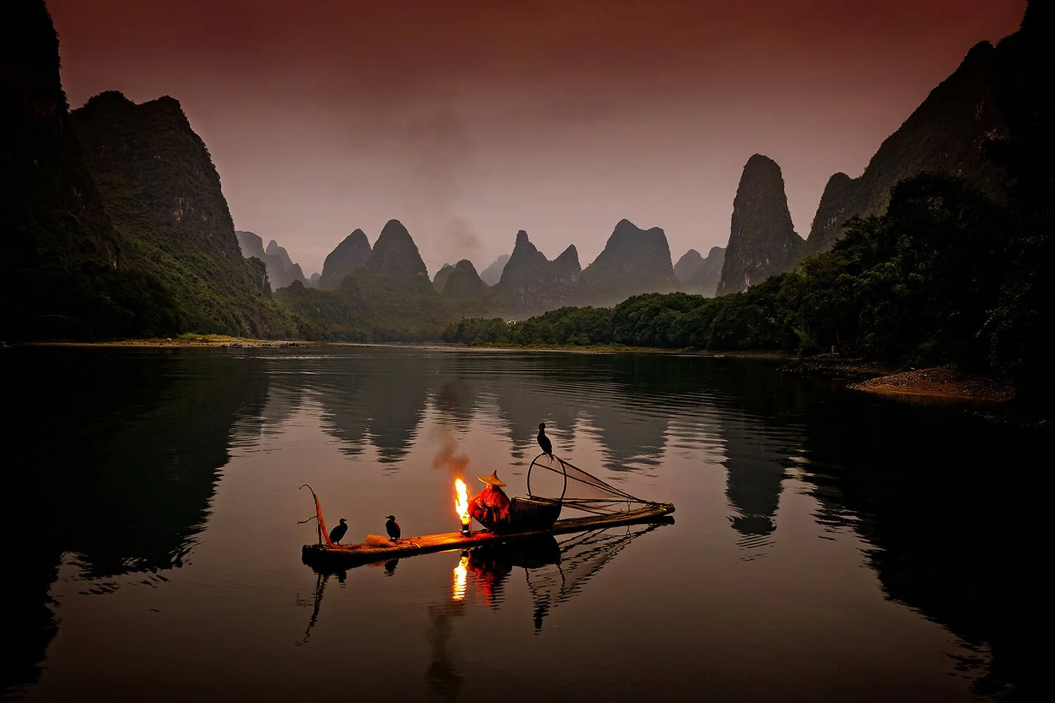 A person wearing a traditional Asian conical hat sitting on a bamboo raft with a small fire, accompanied by three cormorants, floating on a river surrounded by towering limestone karst formations at dusk.
