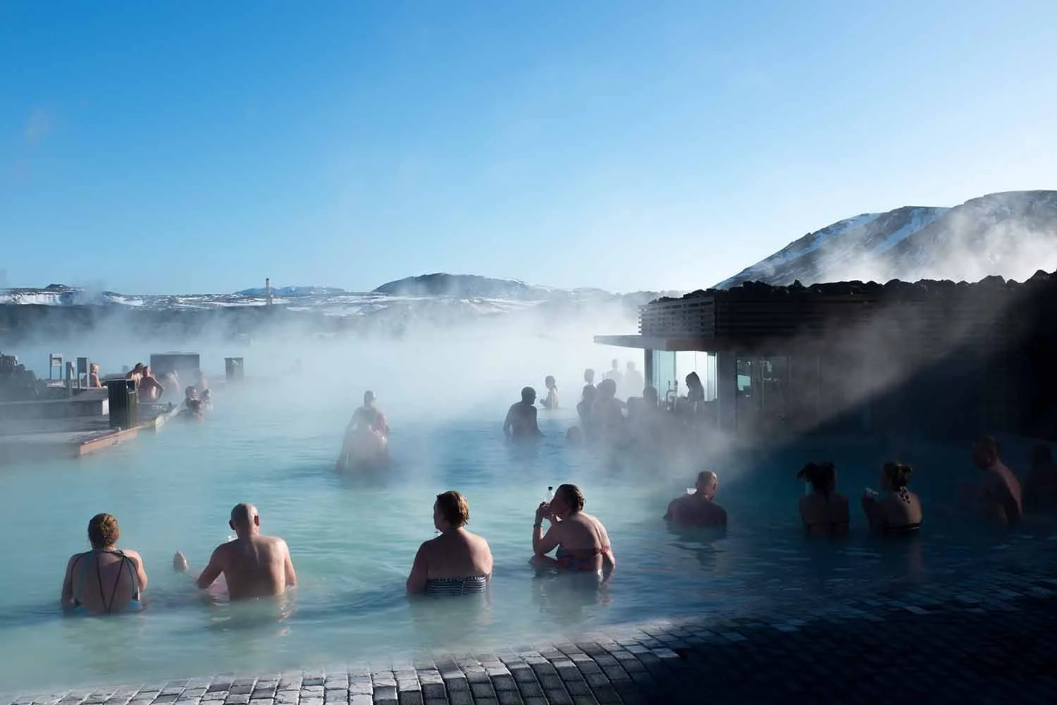People relaxing in an outdoor geothermal hot spring pool with steam rising, snow-covered mountains in the background, and a clear blue sky.