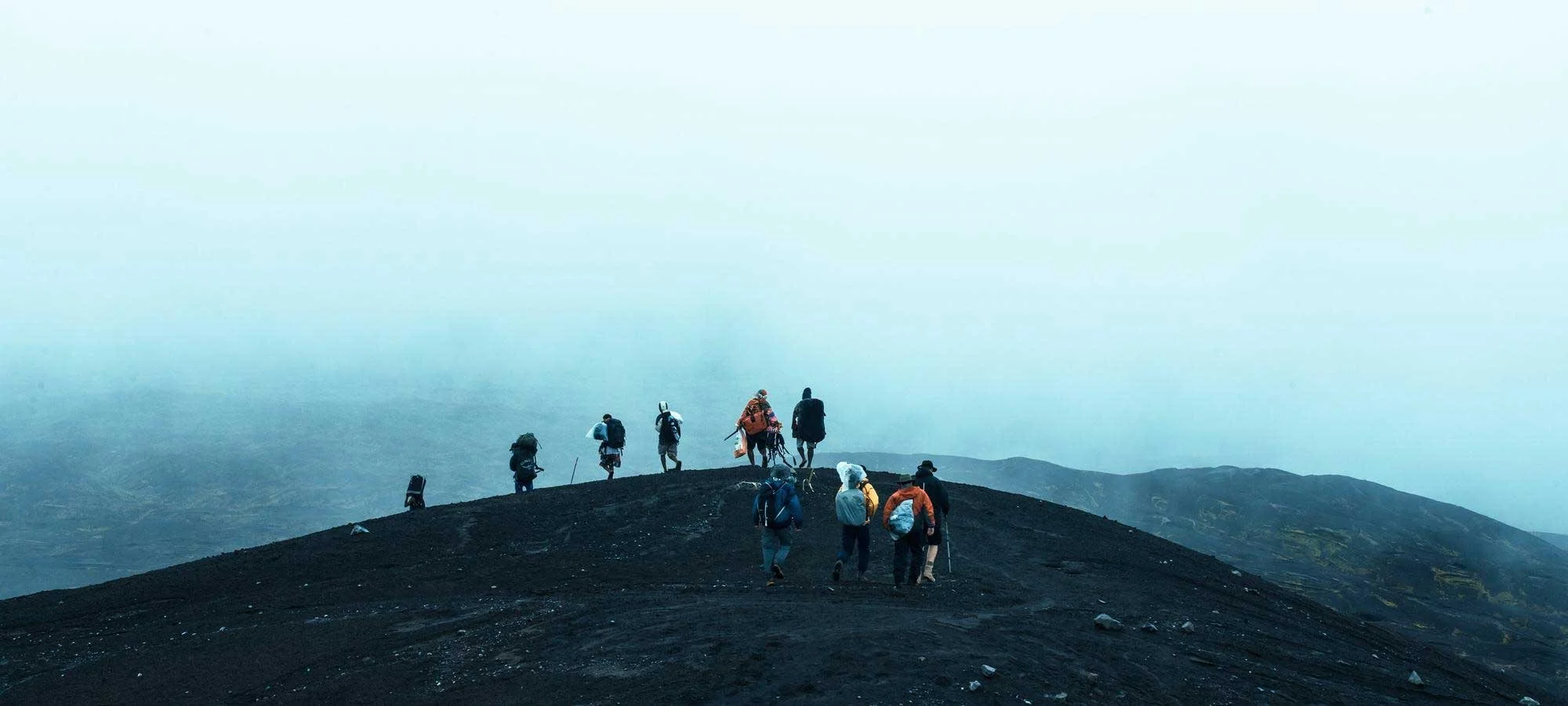 Group of hikers walking on a dark mountain trail with a foggy sky and distant mountains in the background.