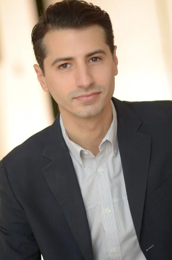 A portrait of a young man with dark hair, wearing a dark suit jacket over a light, checkered dress shirt, smiling subtly, with a soft, blurred background.