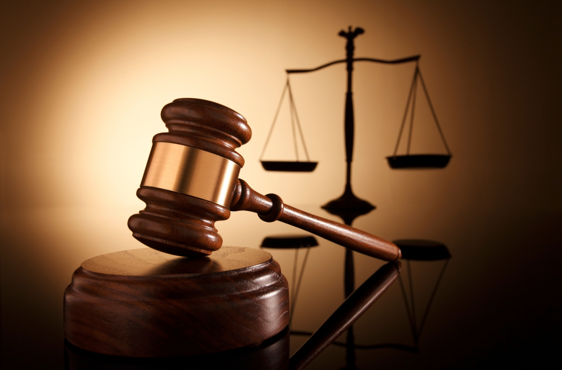 A wooden judge's gavel resting on a sound block with a scale of justice silhouette in the background.