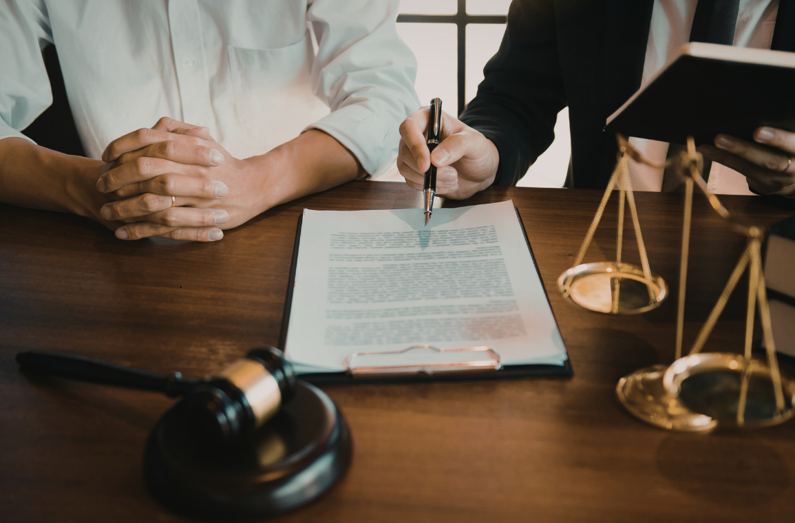 Two professionals signing a legal document at a wooden desk with a gavel, a scale of justice, and a folder nearby.