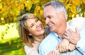 A smiling young woman hugging an older man outdoors amid fall foliage.