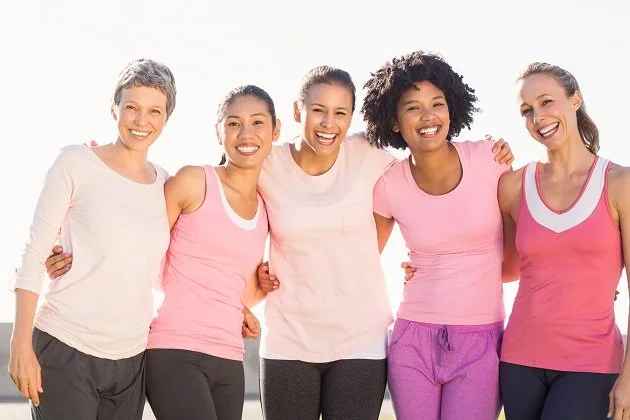 Group of five women smiling and standing close together with arms around each other, wearing workout clothes in pink, white, gray, and purple.