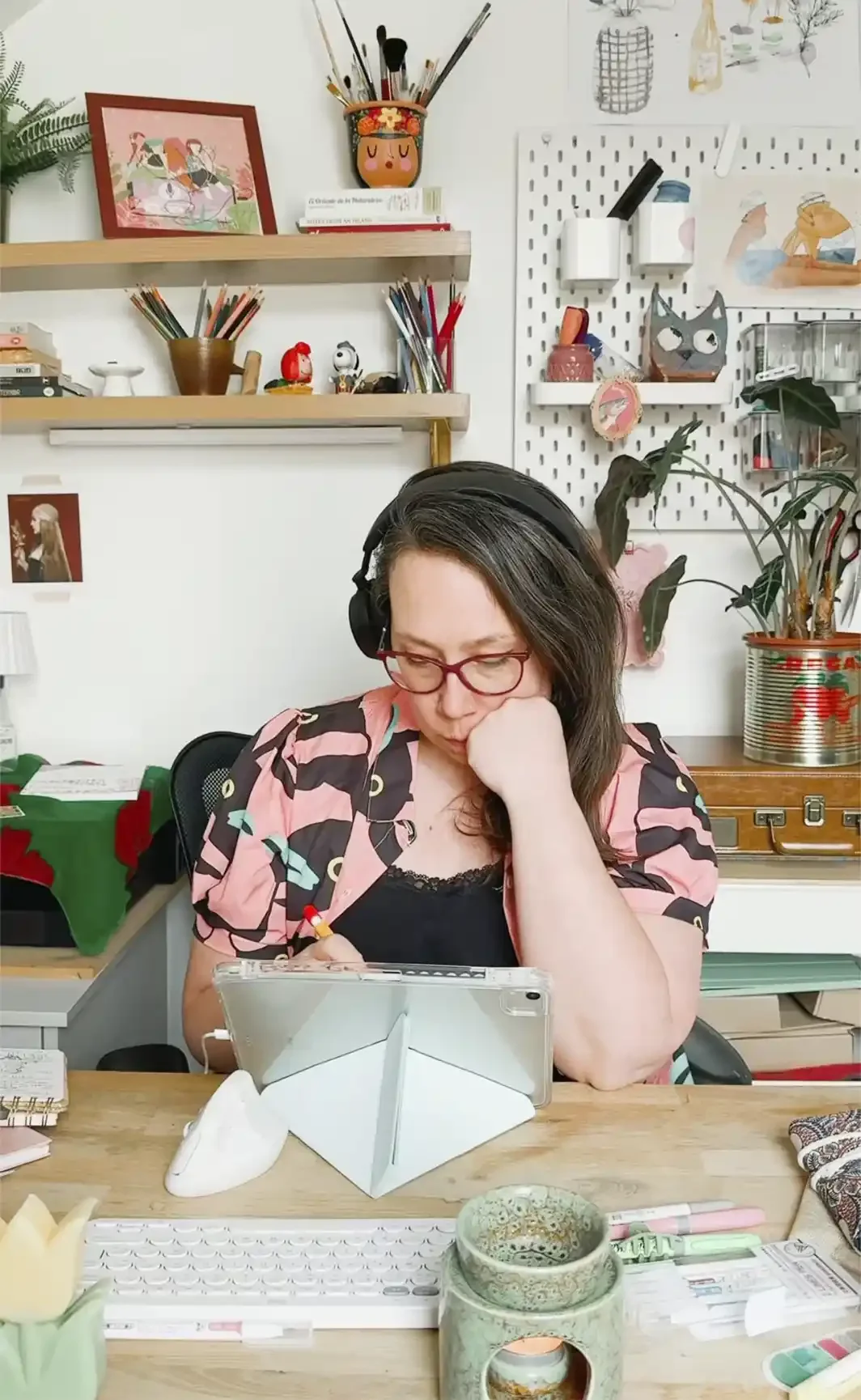 Patricia Corrales Illustration is sitting at a desk, wearing headphones, and looking at a tablet. The background shows shelves with art supplies, plants, and decorative items in a creative workspace.