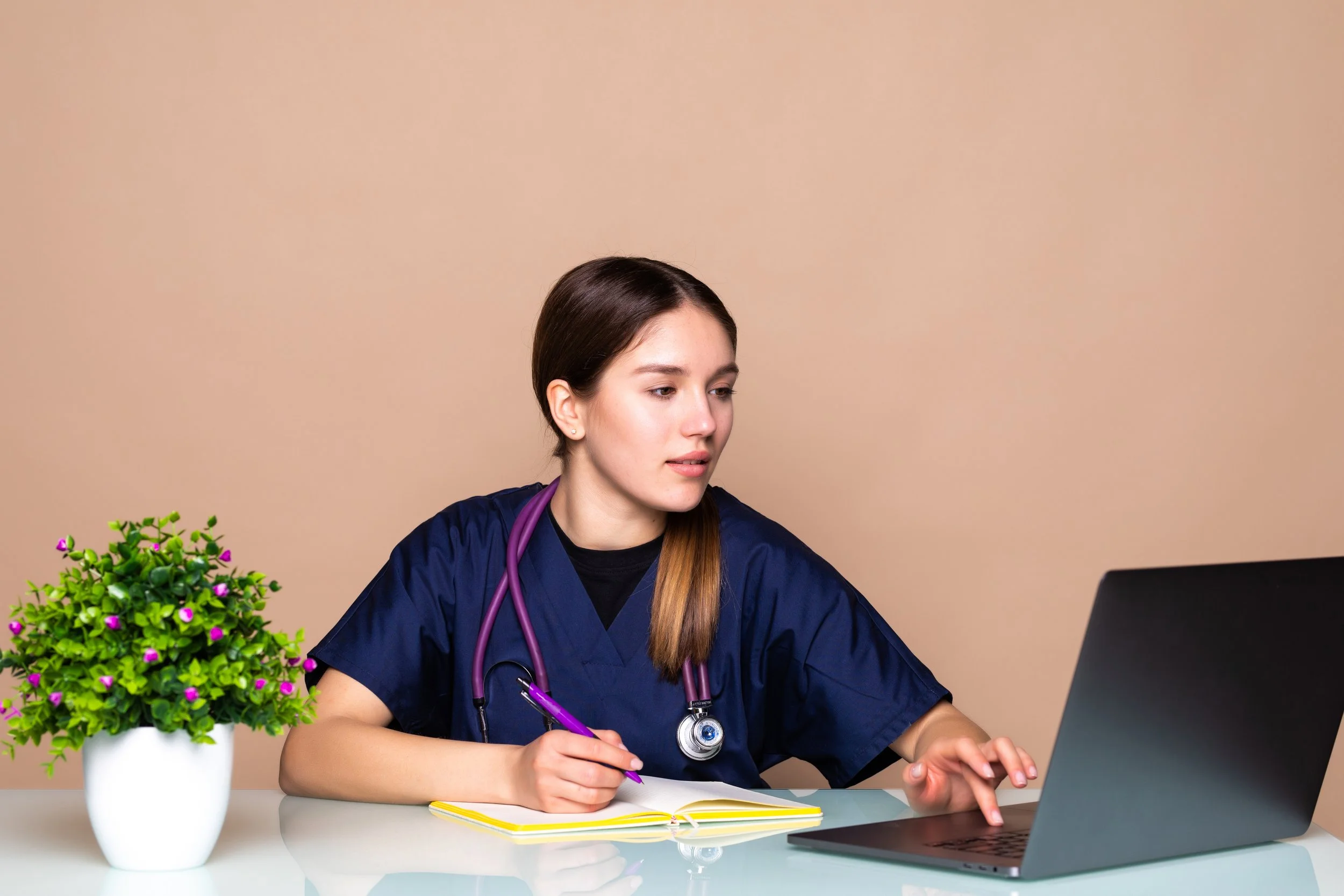 A young female healthcare professional with a stethoscope around her neck, sitting at a desk with a laptop, notebook, and purple pen, looking at the screen. There is a potted plant with small green leaves and purple flowers on her left side.