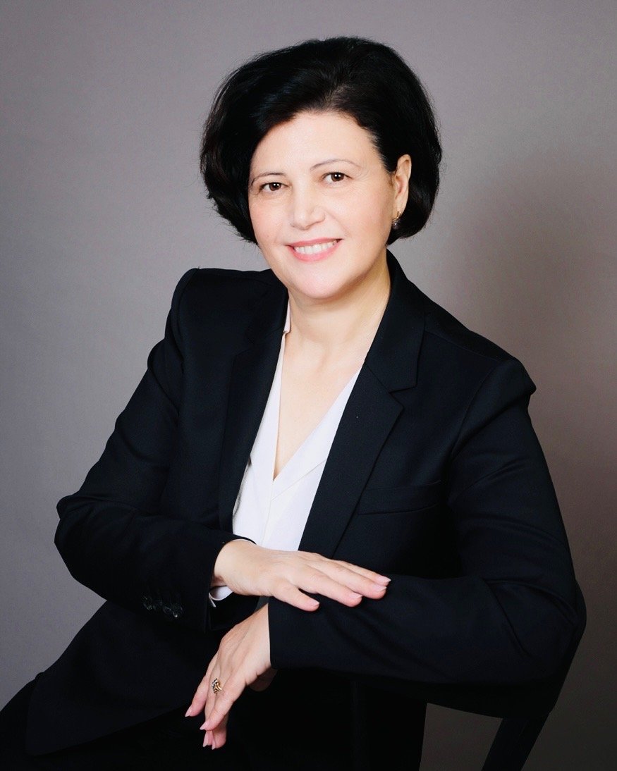 A woman with black hair wearing a black blazer over a white shirt, sitting against a gray background, smiling with her arms resting on a surface.