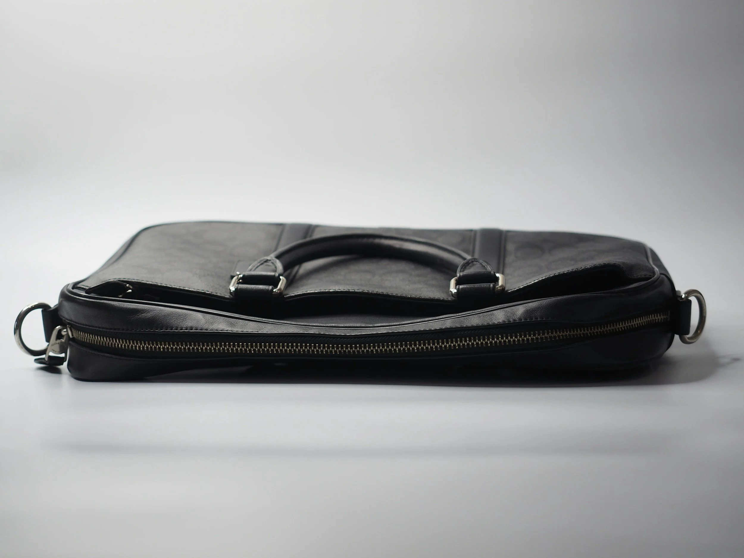 Black leather briefcase with a zipper, placed on a white surface with a plain light gray background.