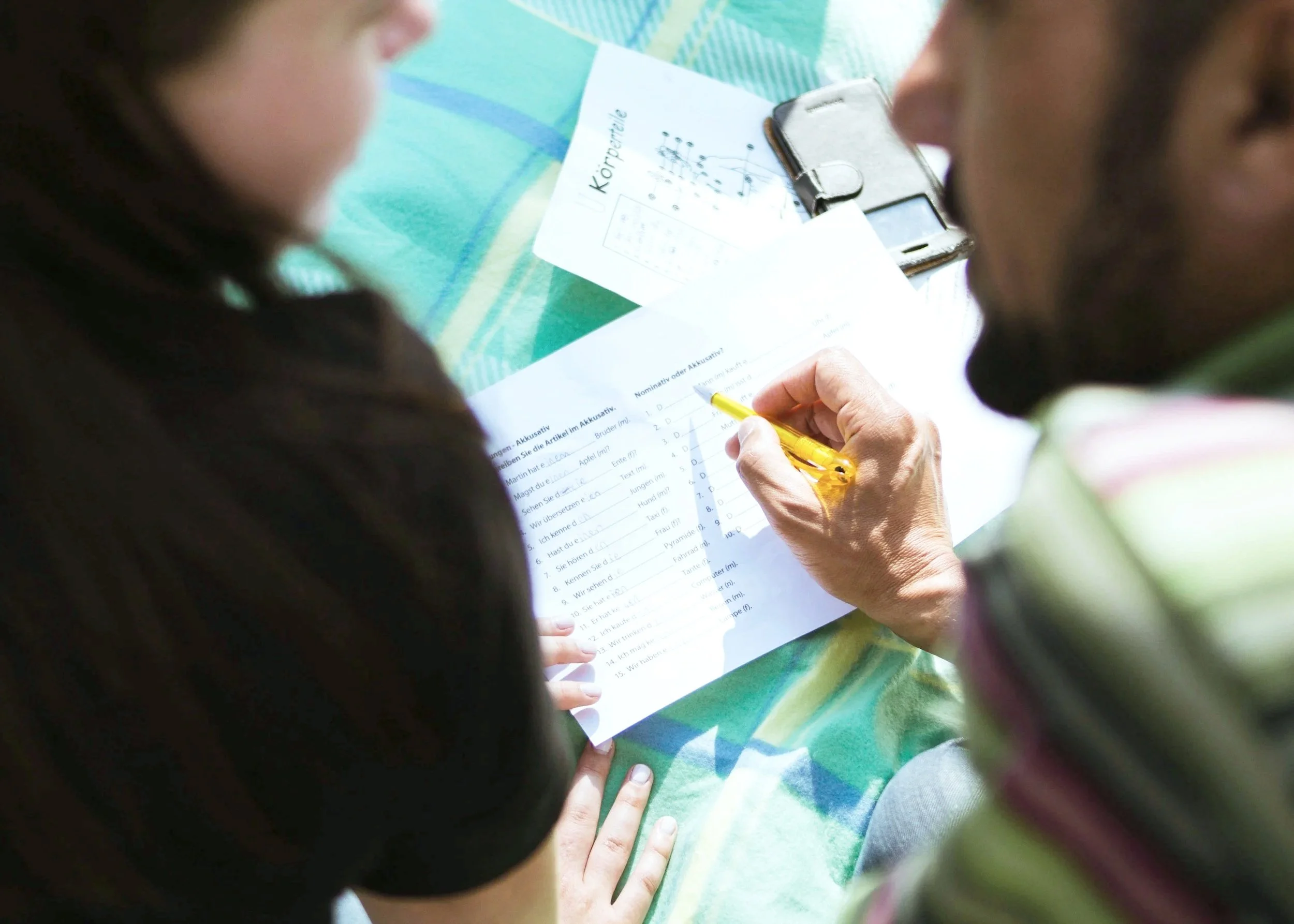 A man and a girl sitting at a table, focusing on a worksheet the man is filling out with a yellow pen. The table has some papers and a phone on it. The worksheet contains German language exercises.