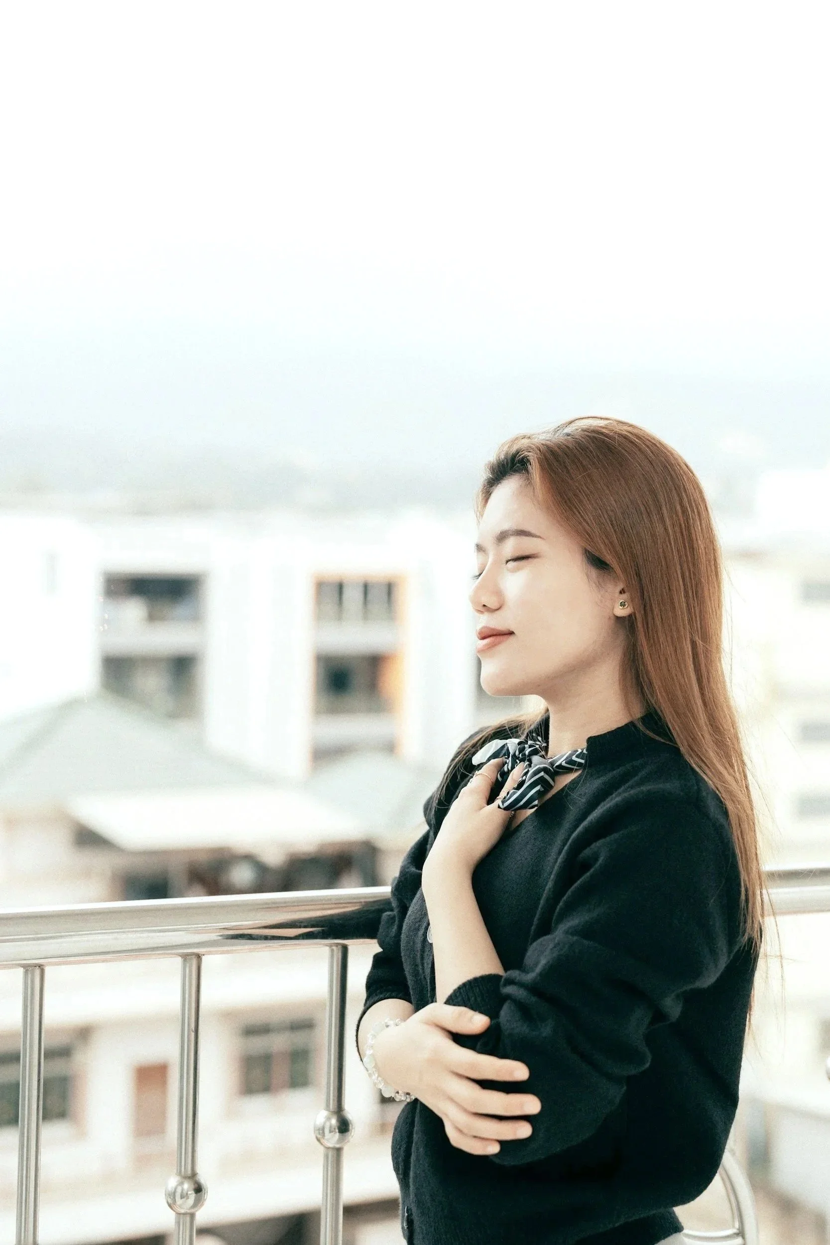 A woman with red hair and closed eyes standing in profile outdoors, leaning on a railing with a cityscape of buildings in the background.