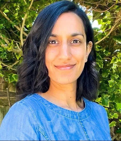 A woman outdoors with green foliage in the background, smiling and wearing a blue top with shoulder-length black hair.
