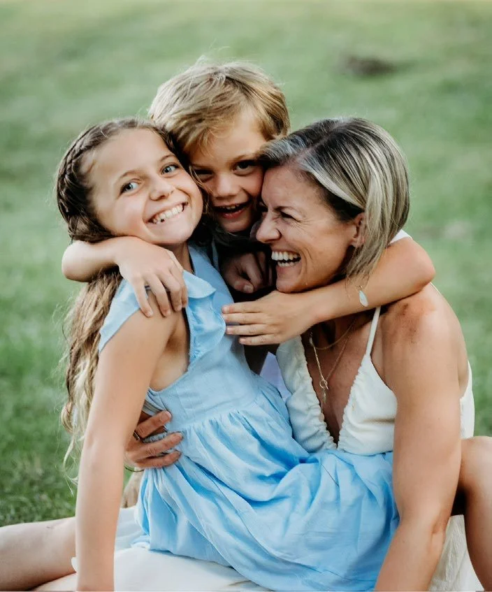 Three people, a woman and two children, hugging and smiling outdoors on grass.