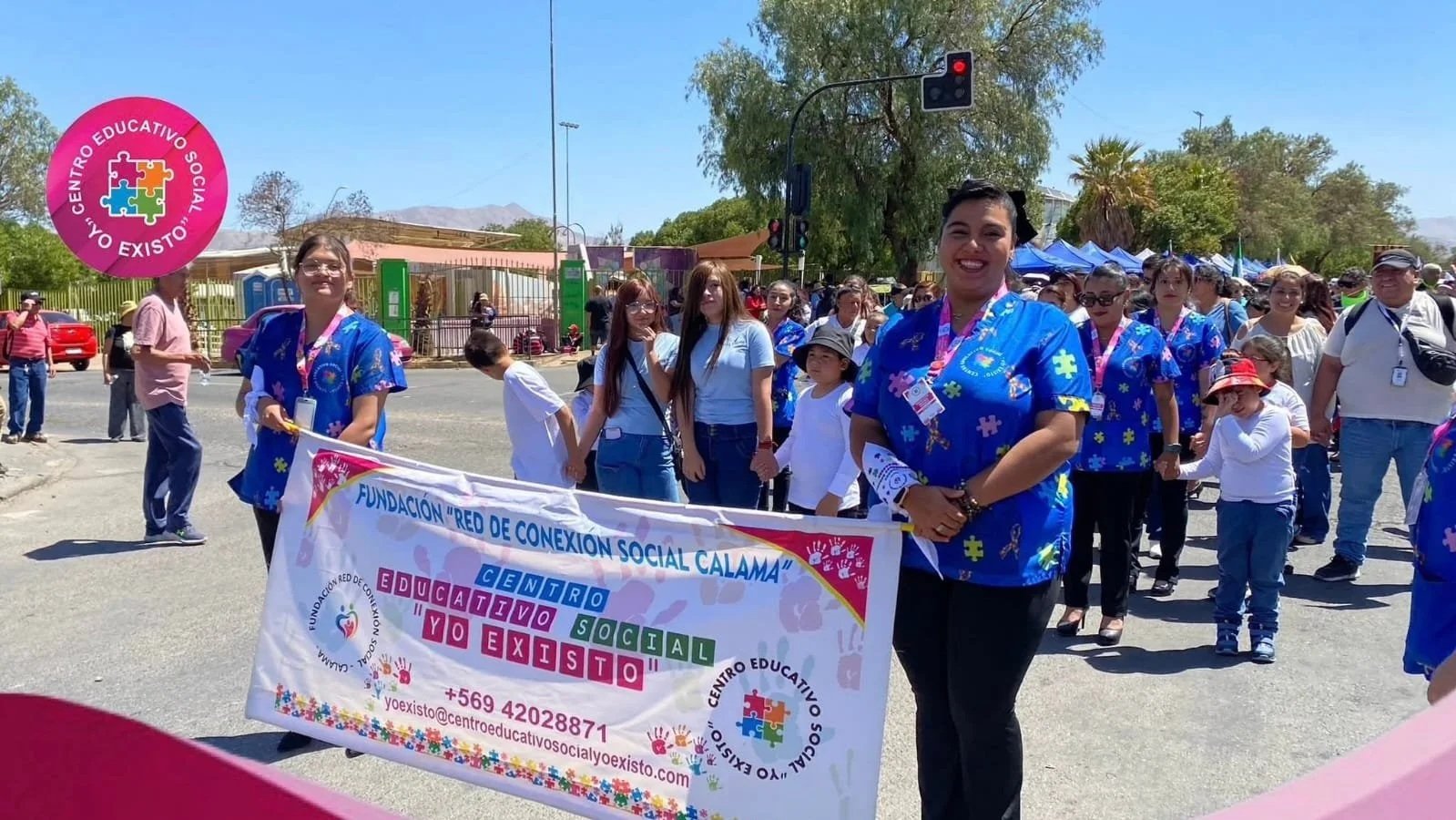 Grupo de personas participando en una marcha o evento, sosteniendo una pancarta que dice 'Fundación Red de Conexión Social Calama' y 'Centro Educativo Social Yo Existo', en un día soleado en las calles de una ciudad con árboles y edificios en el fondo.