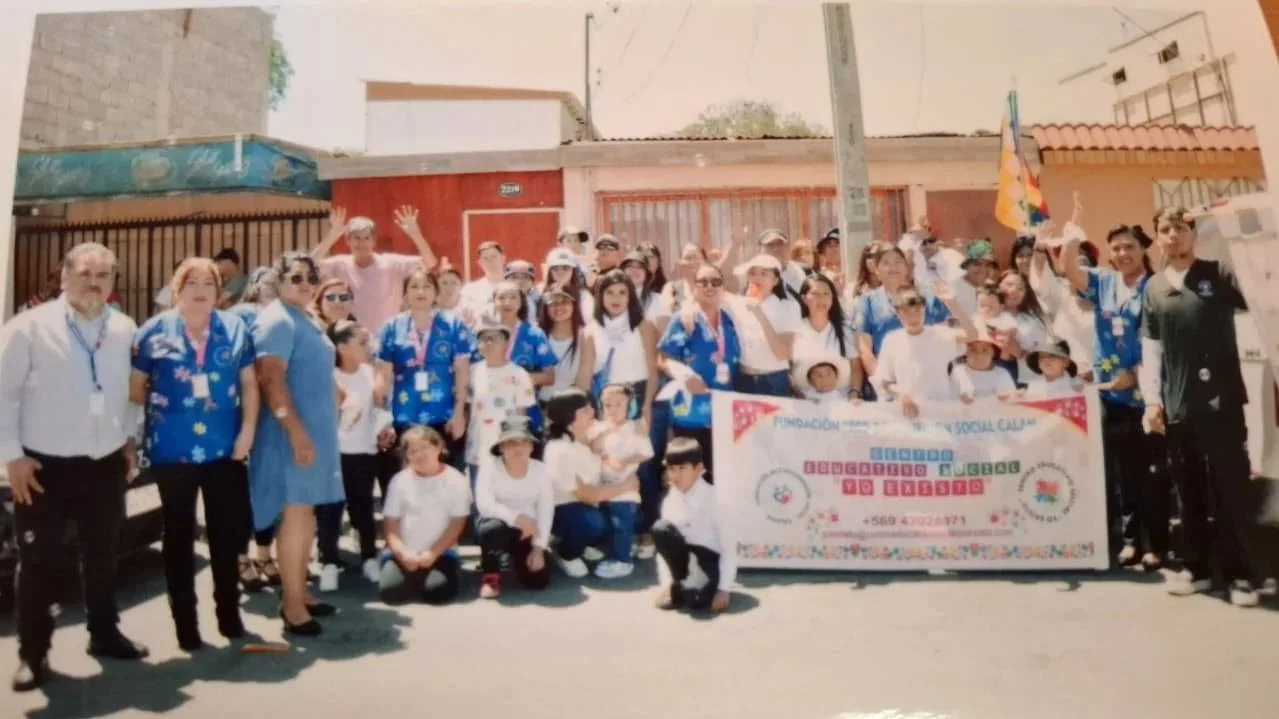 Grupo de personas con cartel en un evento comunitario, algunos llevan camisetas azules y sombreros, en una calle con edificios de fondo.