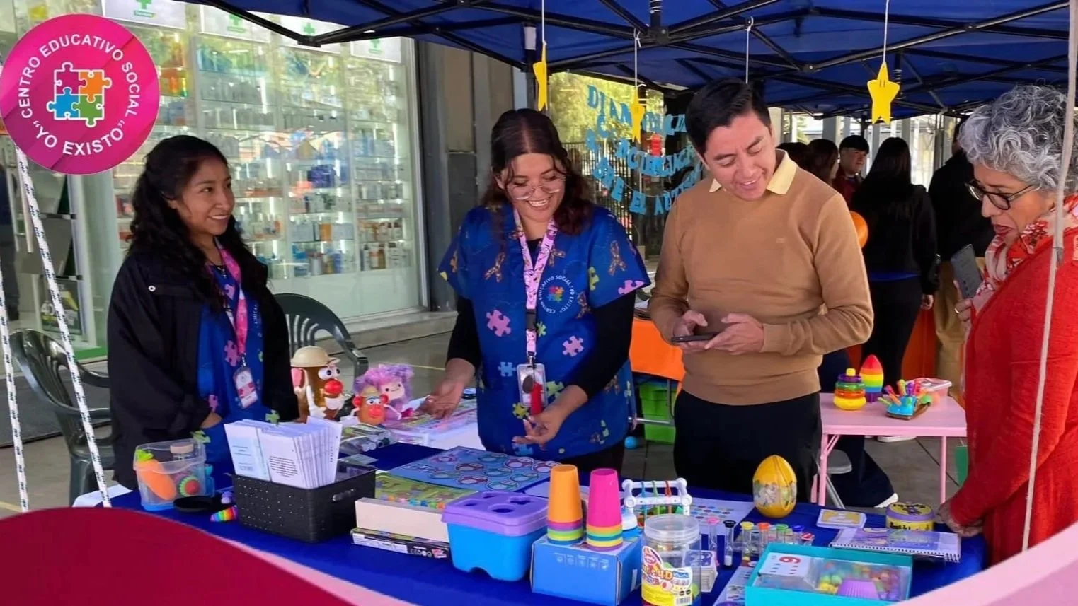 Personas en una feria de ventas con artículos de actividades para niños, decoraciones y folletos, en un espacio cubierto con toldos azules y varios asistentes observando y comprando productos.