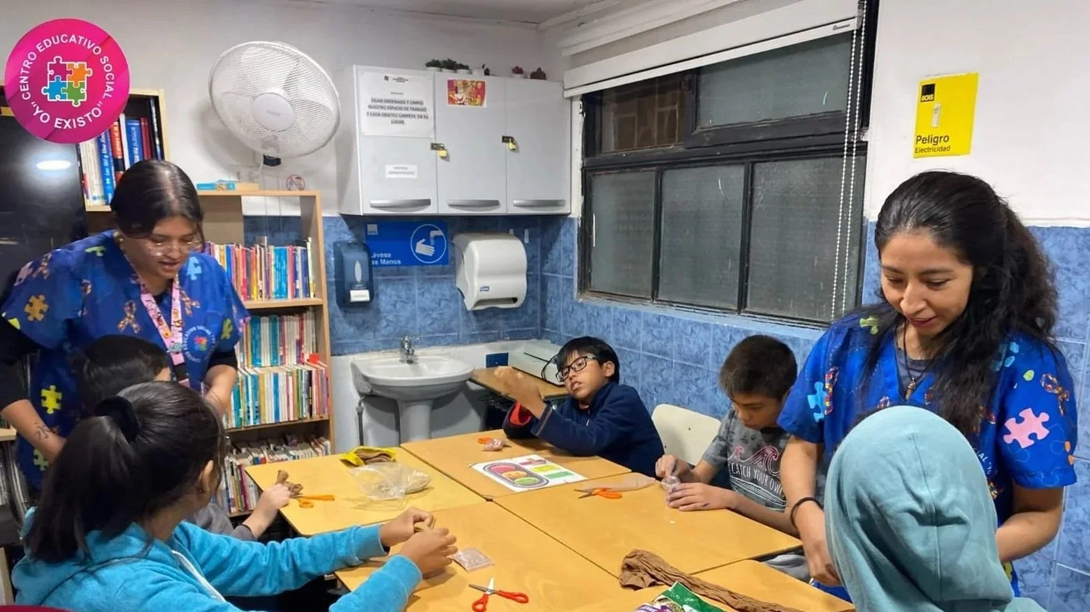Niños en una clase de terapia ocupacional con dos adultos, en una habitación con libros y pisos azules, realizando actividades manuales.