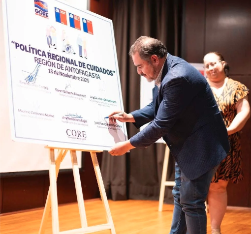 Hombre firmando un documento en un evento oficial, con una mujer observando, en un escenario con cortinas oscuras y un cartel en un caballete.