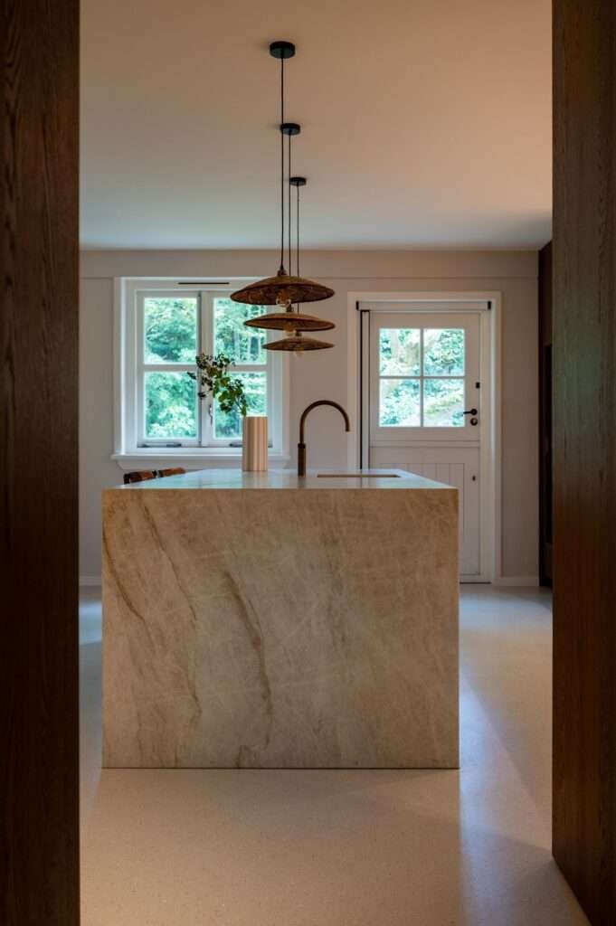 Intérieur moderne d'une cuisine avec un îlot en pierre, un robinet noir, une suspension en bois, fenêtre avec vue sur la nature et porte en bois blanc
