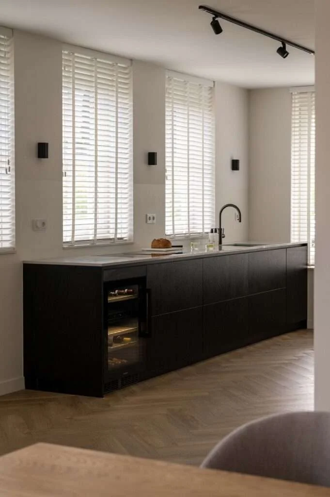 Cuisine moderne avec plan de travail blanc, meuble noir, évier, robinet, et fenêtres avec stores en bois, sols en bois clair.