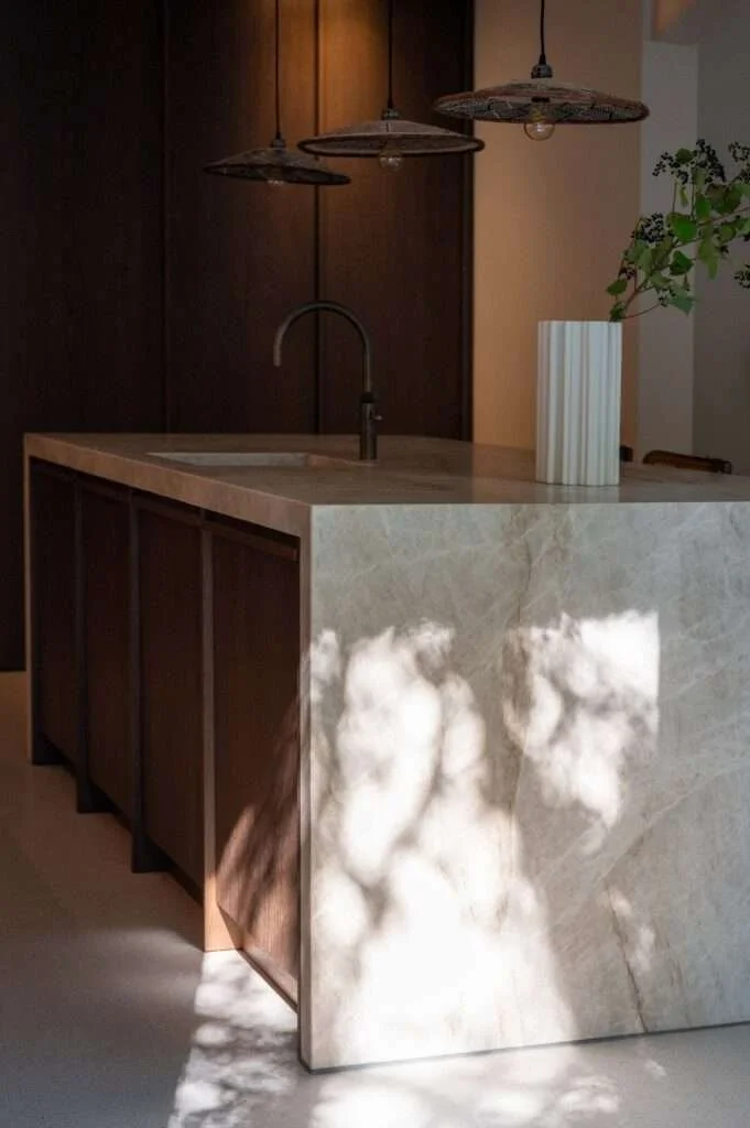 Une cuisine moderne avec un îlot en marbre clair, éclairée par trois lampes en rotin suspendues, et décorée avec une vase blanche et un mur en bois foncé.