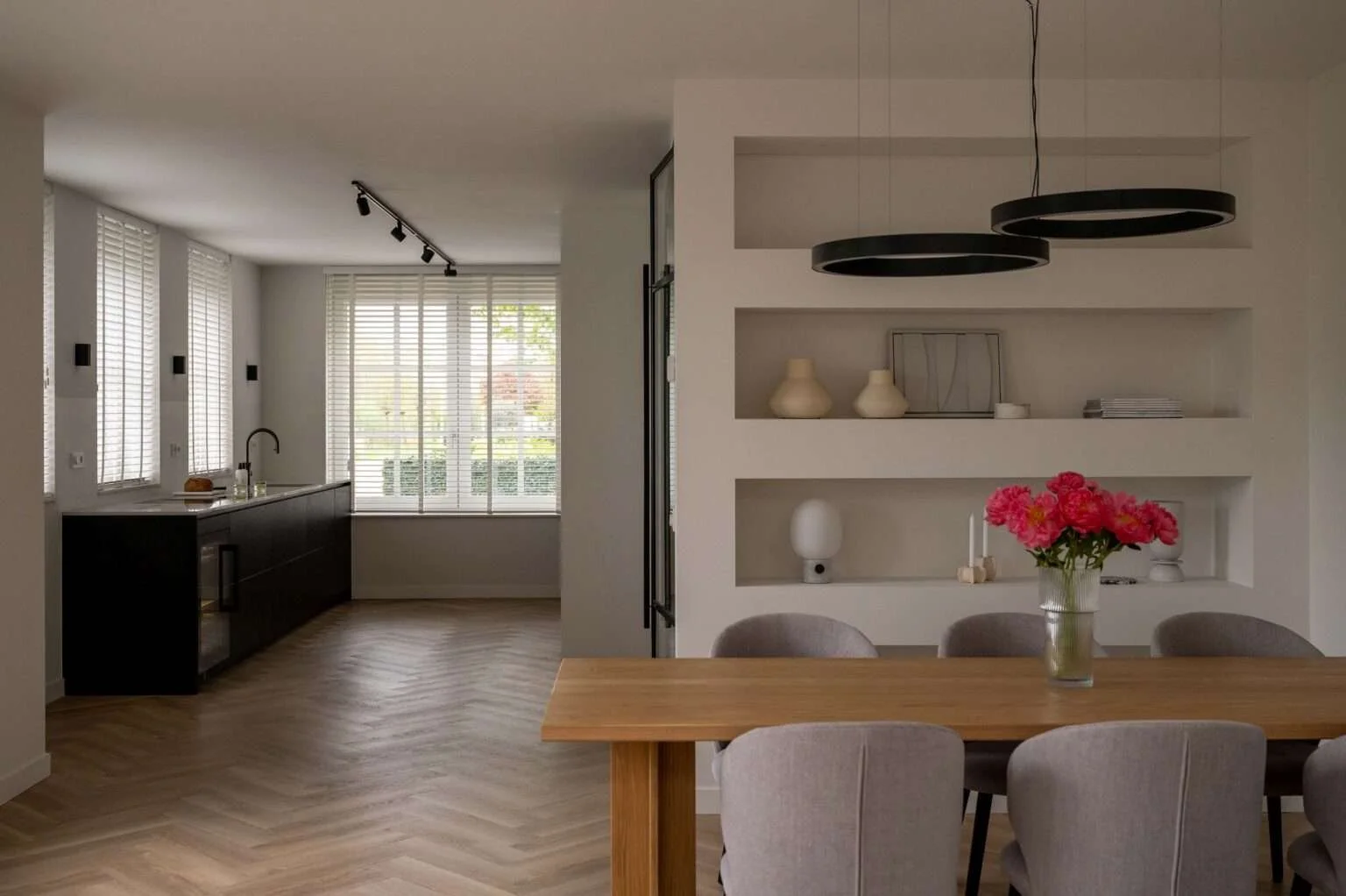 Salle à manger moderne avec vase de fleurs roses sur la table, étagères blanches avec vases et cadres, cuisine en arrière-plan avec fenêtres à volets