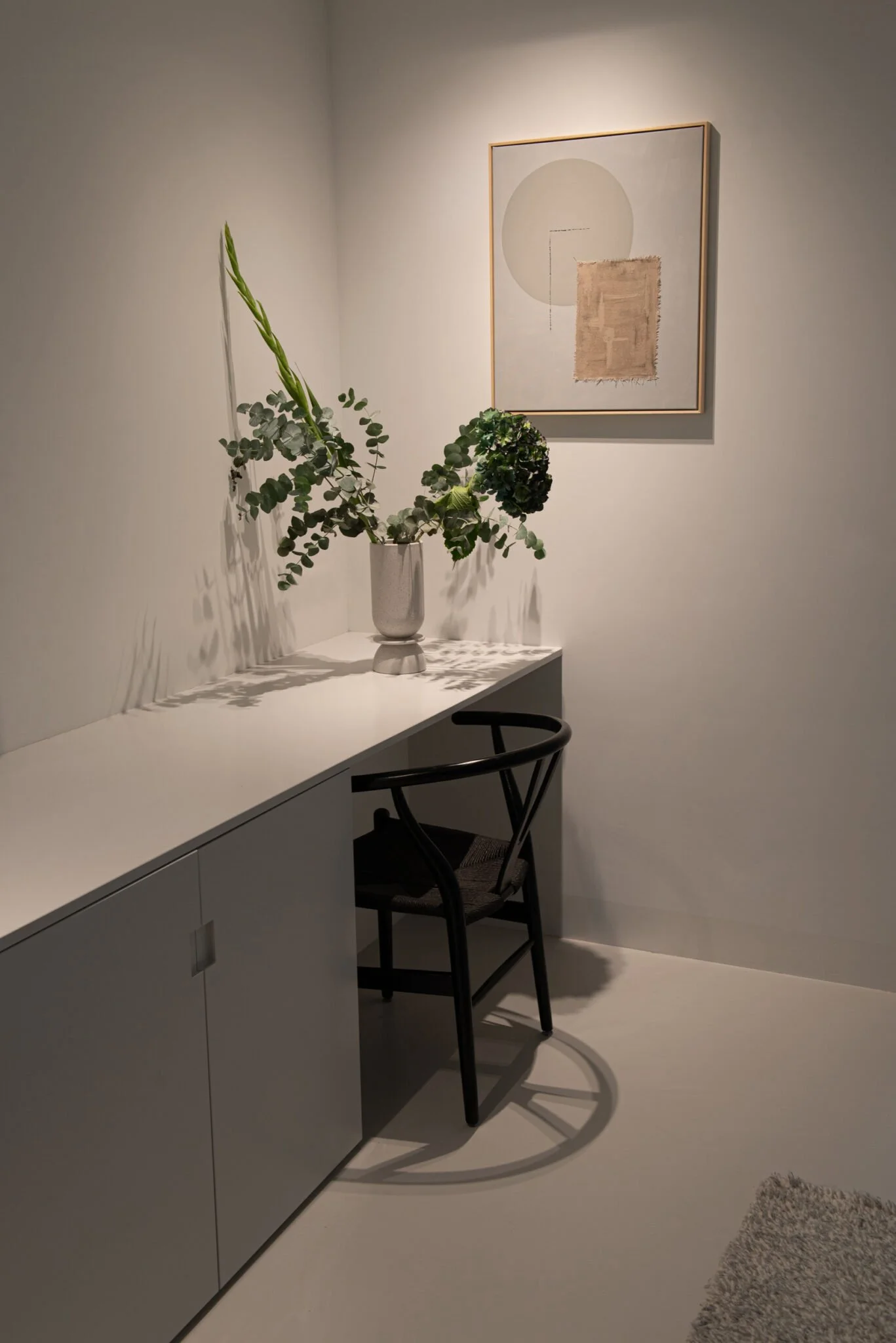 Une pièce minimaliste avec un bureau blanc, une chaise noire, un vase avec des plantes vertes, un tableau abstrait au mur.