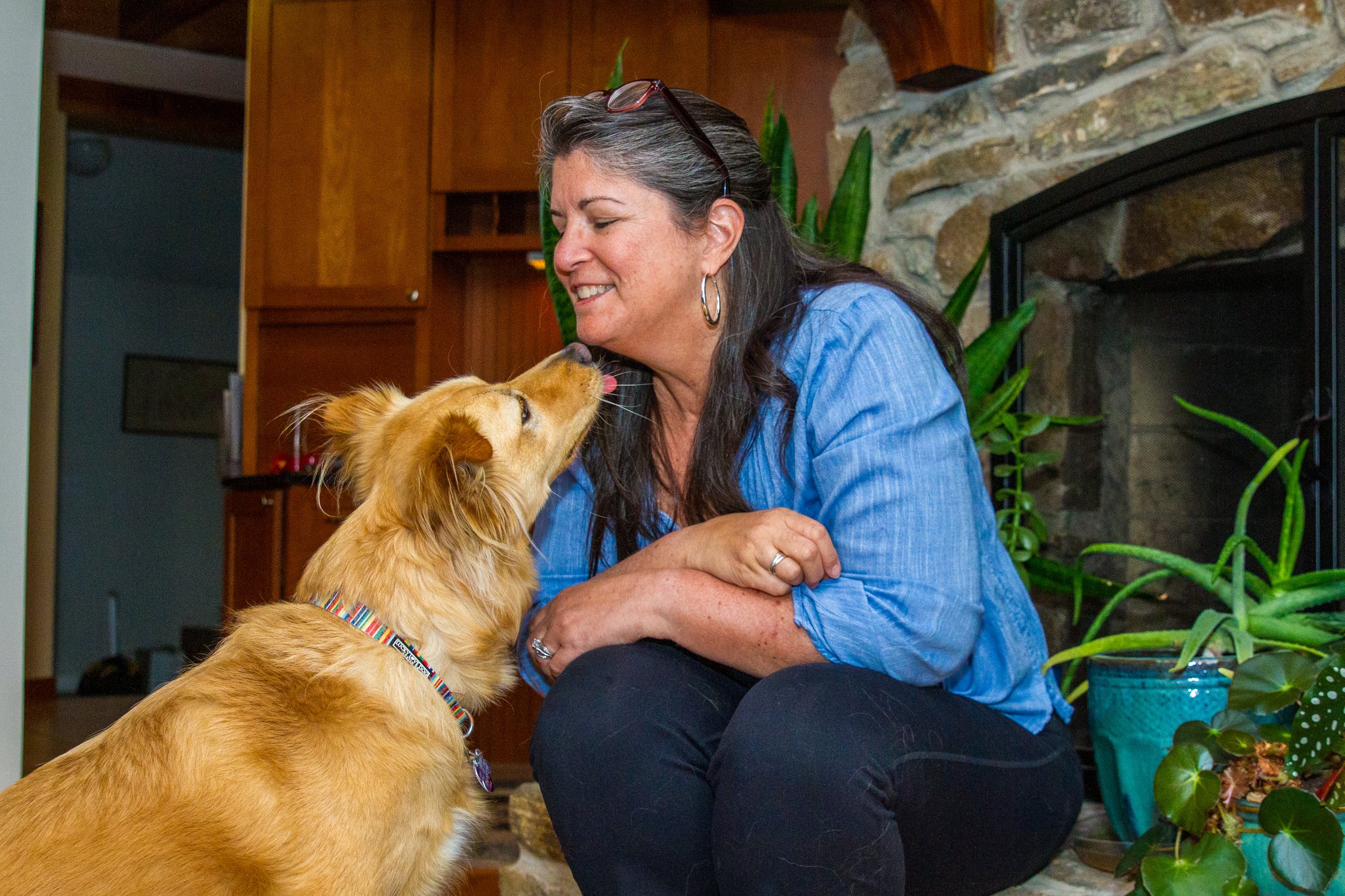 A woman smiling and leaning towards her golden retriever dog indoors by a fireplace, with plants nearby.