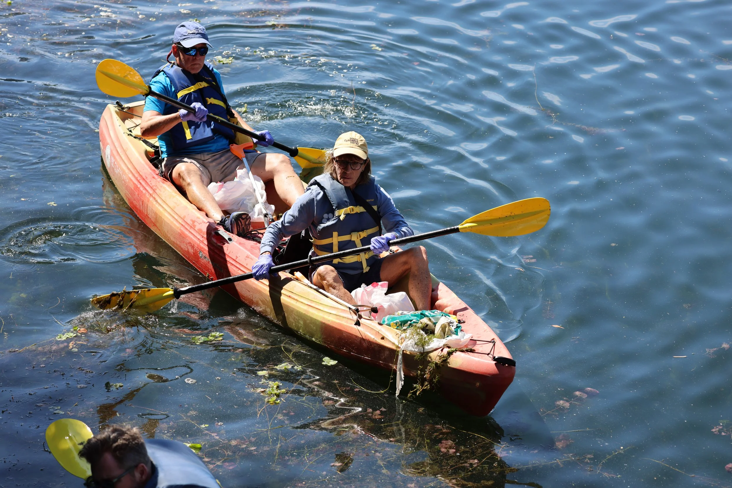 Two people are paddling a tandem kayak on a body of water with some floating debris and plants, wearing life jackets, hats, and sunglasses.
