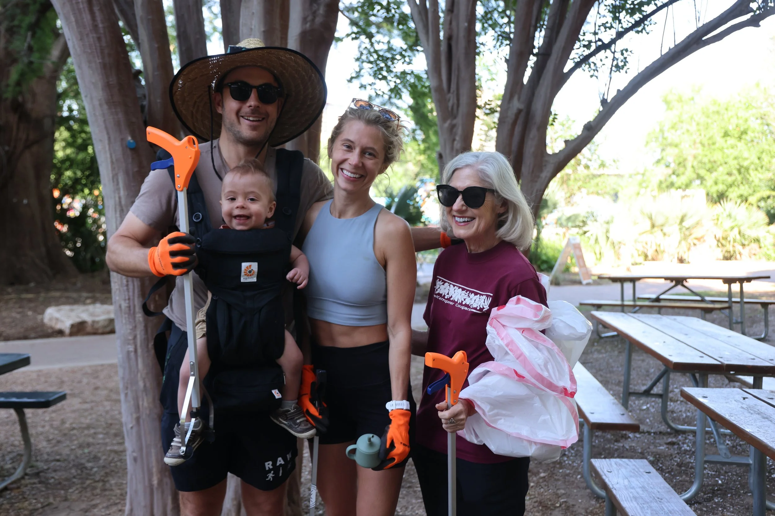 A group of four happy people, including a baby, enjoying an outdoor activity with gardening tools, standing in a park with trees and picnic tables in the background.