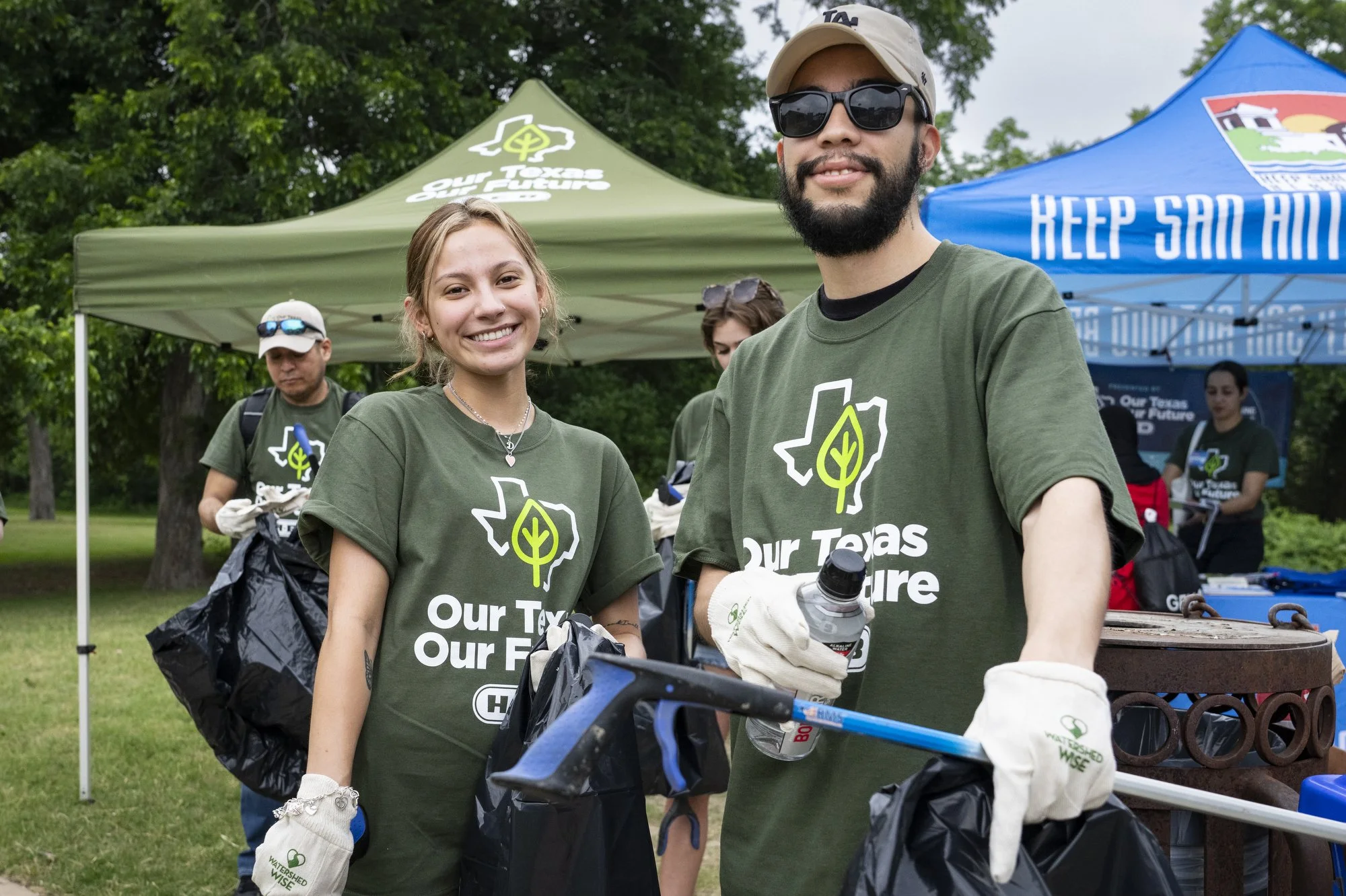 Group of volunteers at an outdoor environmental cleanup, wearing green shirts with Texas outline and tree logo, with tents in the background.