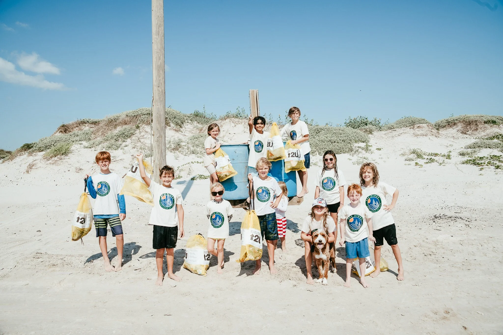 Children on a beach holding bags, with a dog, posing near a blue trash bin and a utility pole, sand dunes in the background on a sunny day.