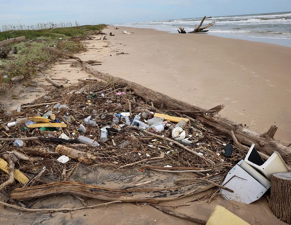 Beach with scattered trash and driftwood along the shoreline, ocean waves in the background, cloudy sky.